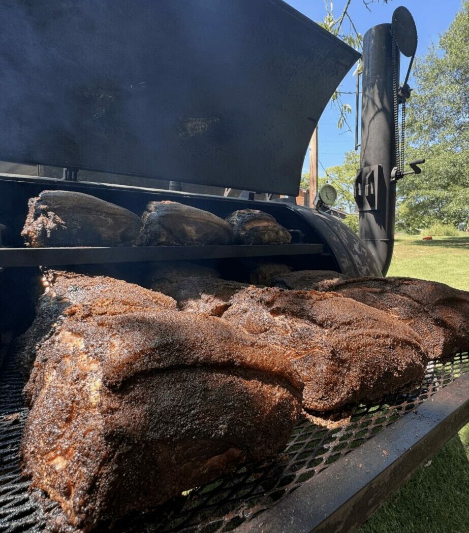 Barbecue grill with cooked meat and a smoker in the background outdoors, trees and blue sky visible.