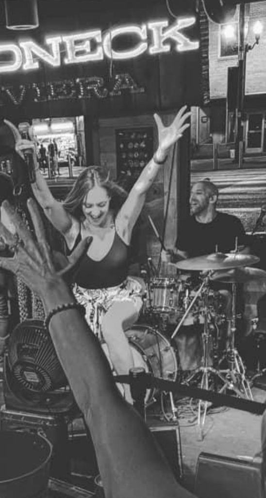 A woman with arms raised performing on stage with a drummer in the background, neon sign and city street visible.