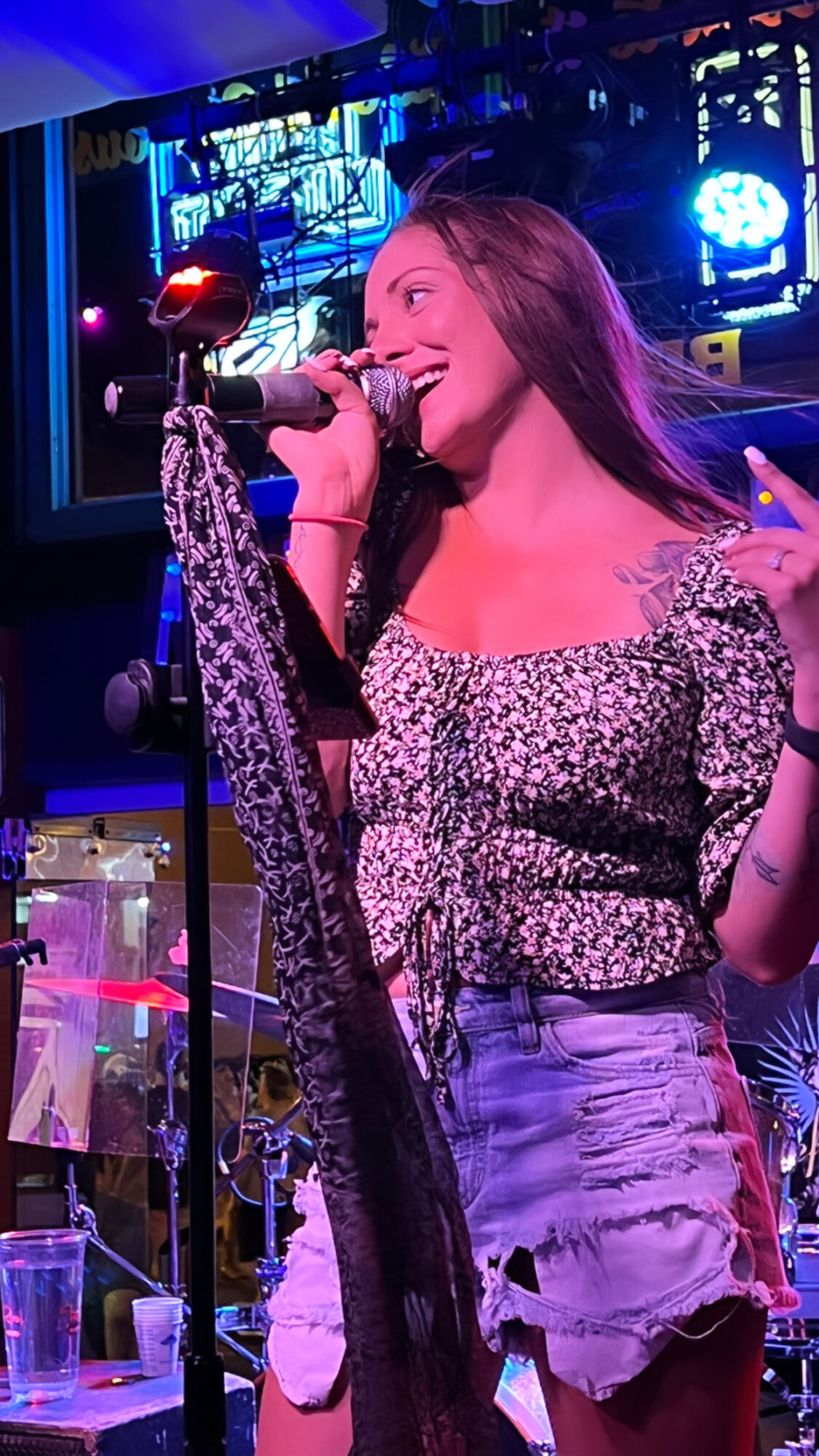 Young woman singing into a microphone on stage with colorful lights, wearing a patterned top and ripped jeans.