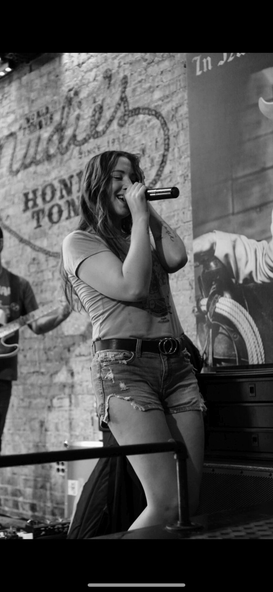 Woman singing into microphone on stage with a guitar in background, black and white image.