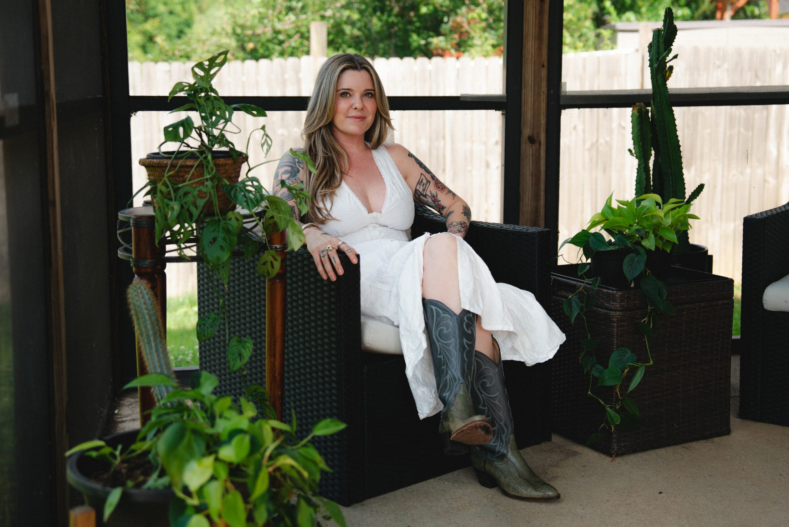 Woman sitting on a black sofa surrounded by potted plants, with a wooden fence and trees outside window