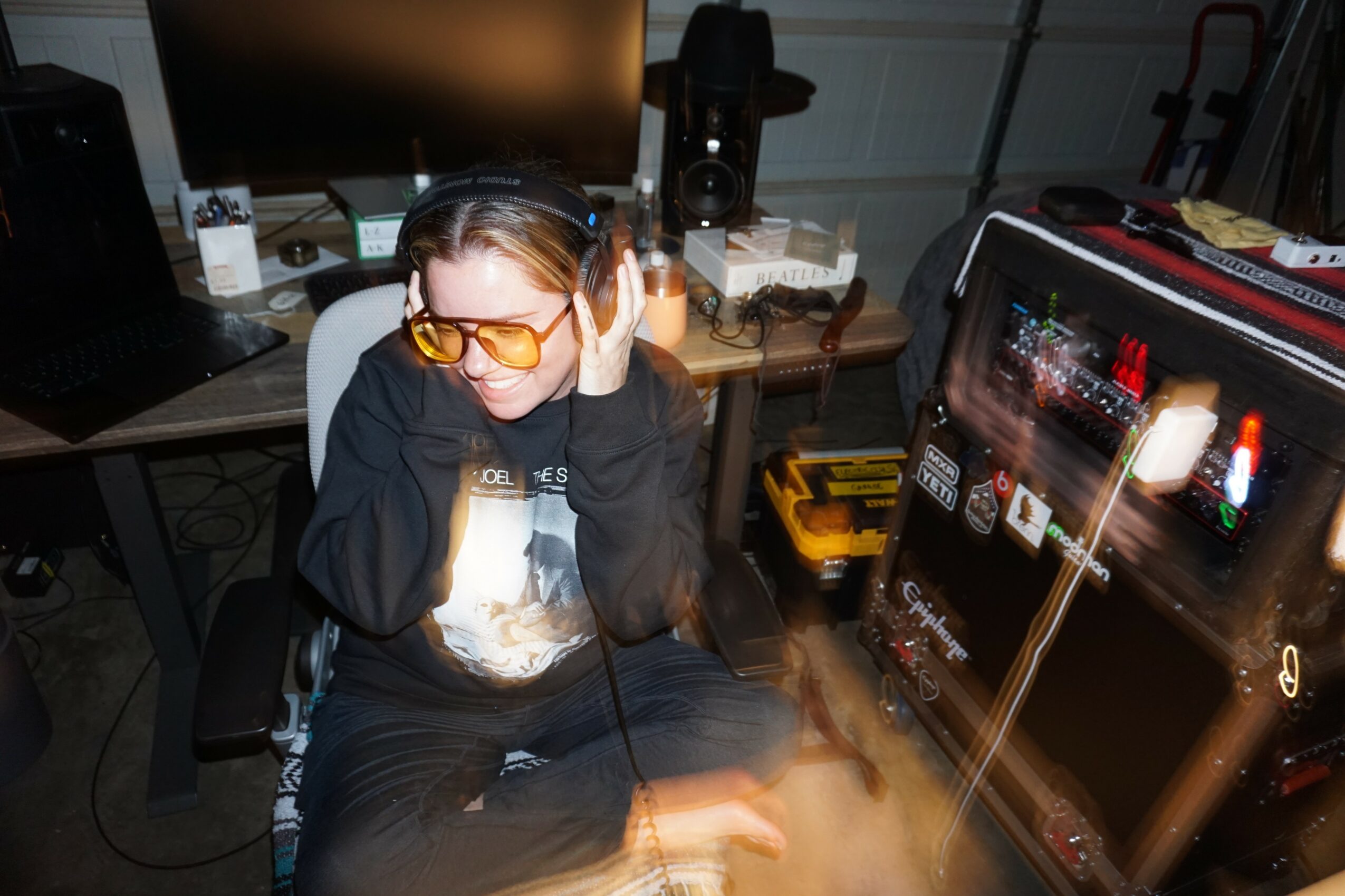 Person wearing headphones and yellow glasses sitting at a desk with a computer and audio equipment, in a room with various electronic devices.