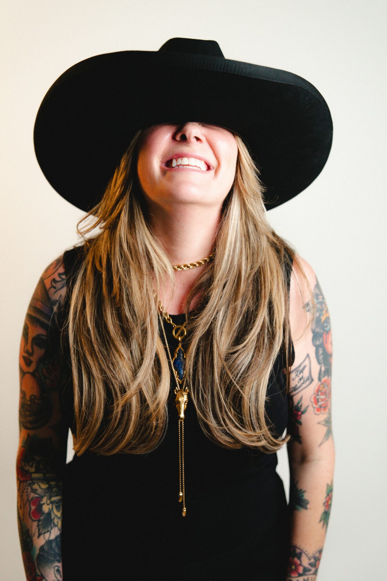 Woman with long hair wearing a large black hat, smiling, and dressed in black with tattoos on arms.