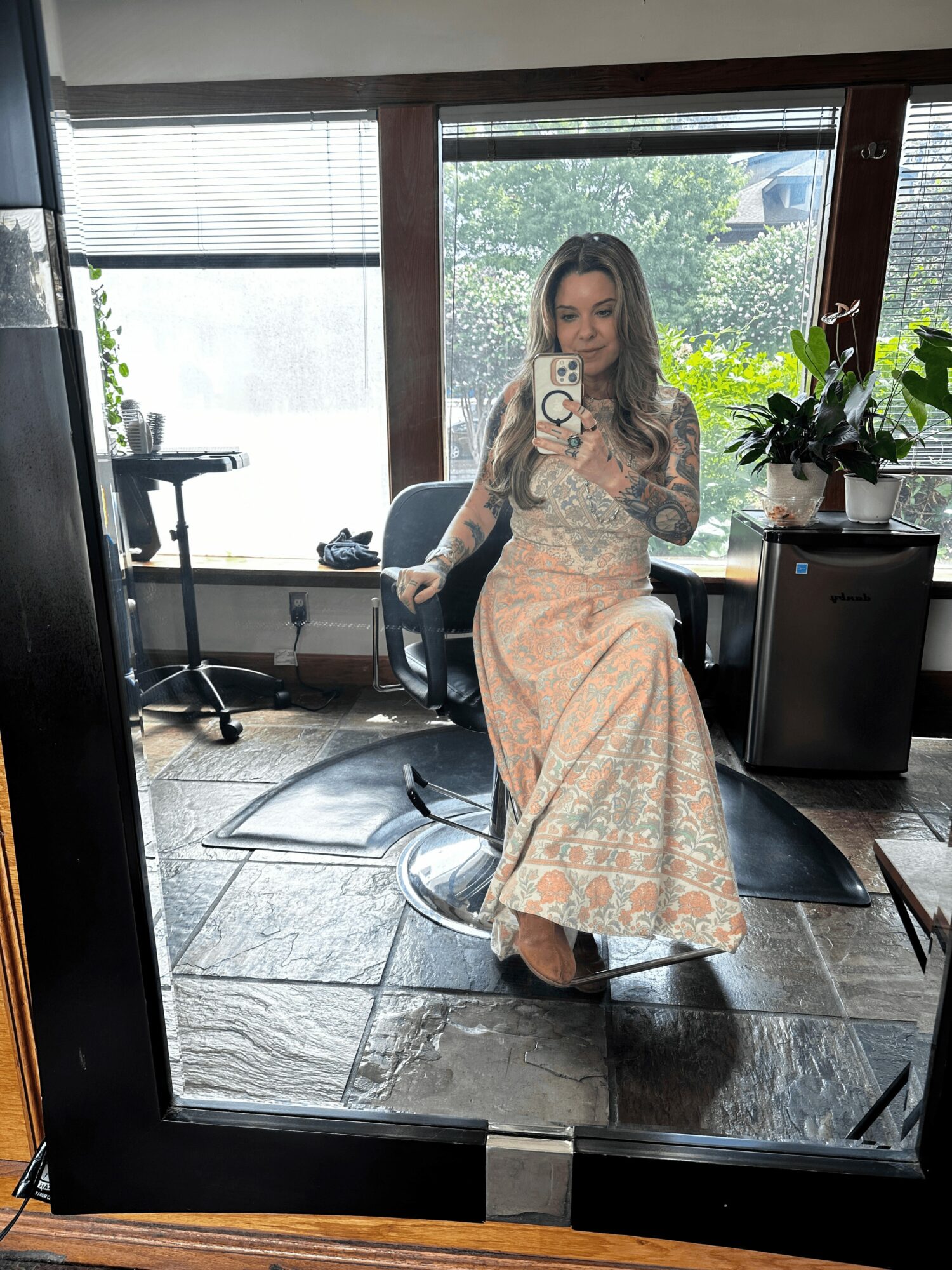 Woman in a long, patterned dress sitting on a chair, taking a selfie in a room with large windows and plants.