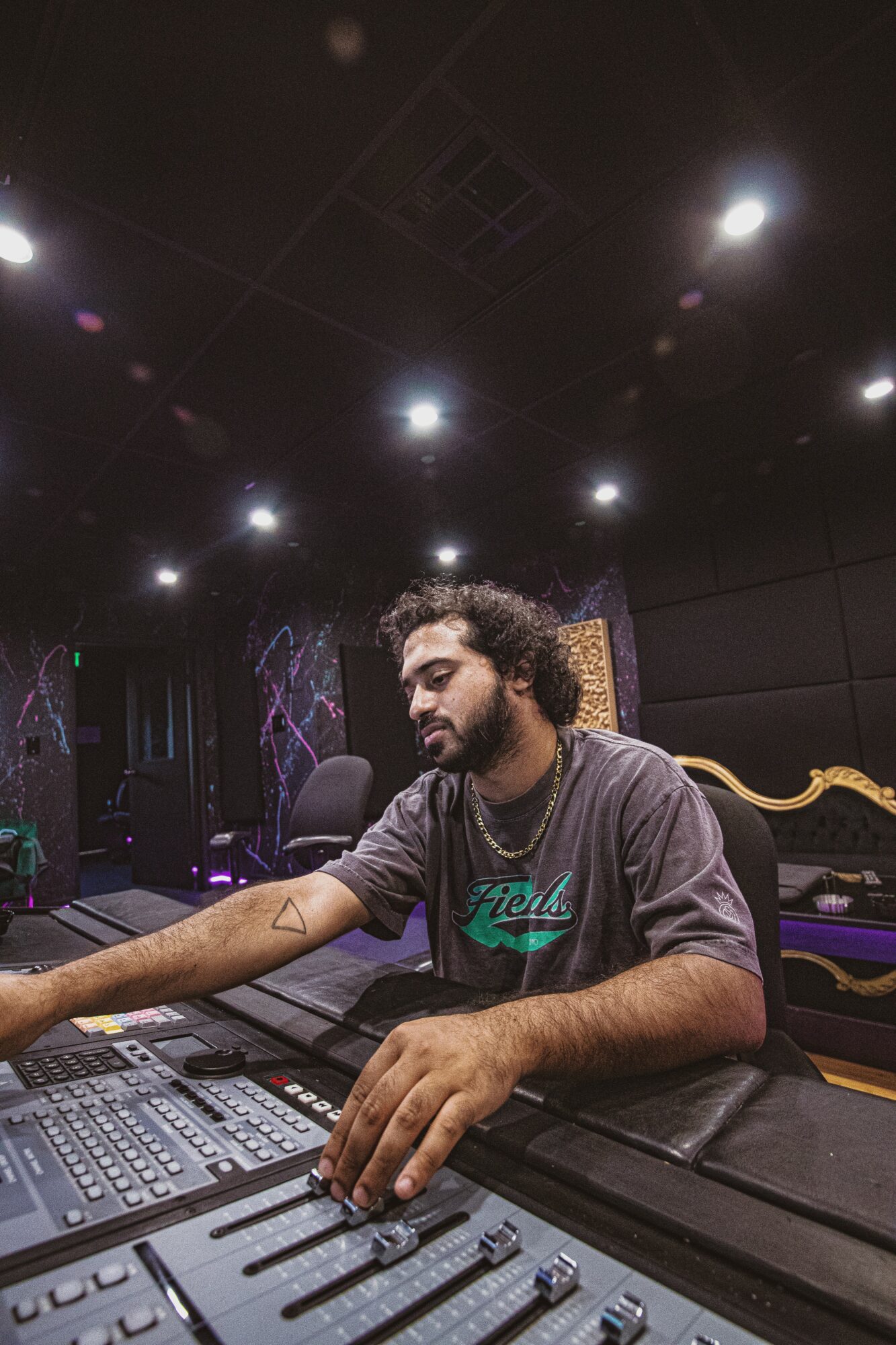 Person with curly hair and beard working at a sound mixing console in a dimly lit room with colorful wall art.