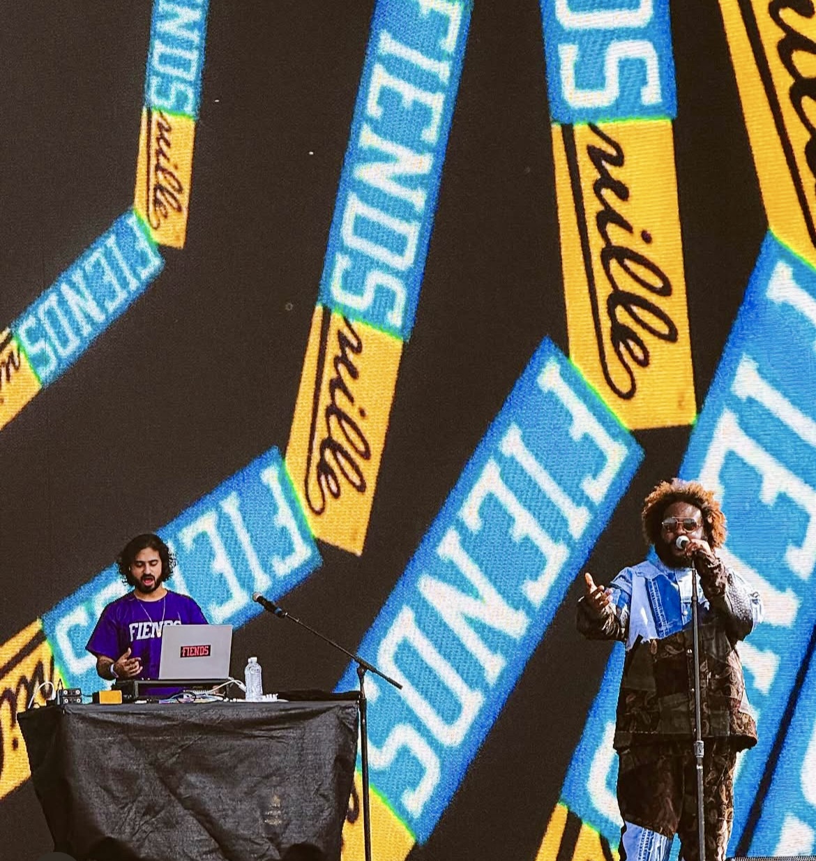Two performers on stage with large colorful banners in the background, one with a laptop and the other singing into a microphone.