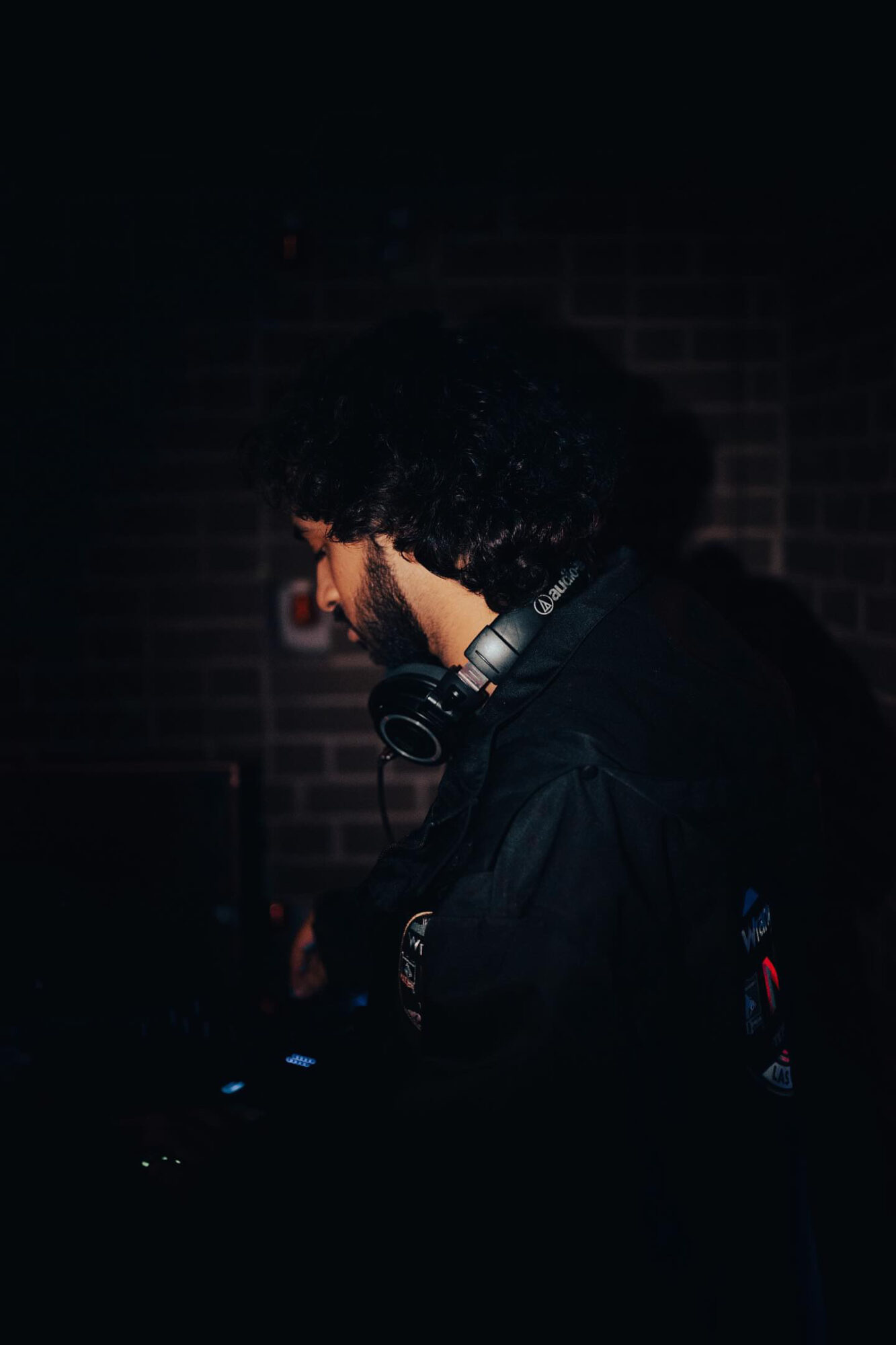 Person with curly hair wearing headphones, facing left in a dark environment with a brick wall background.