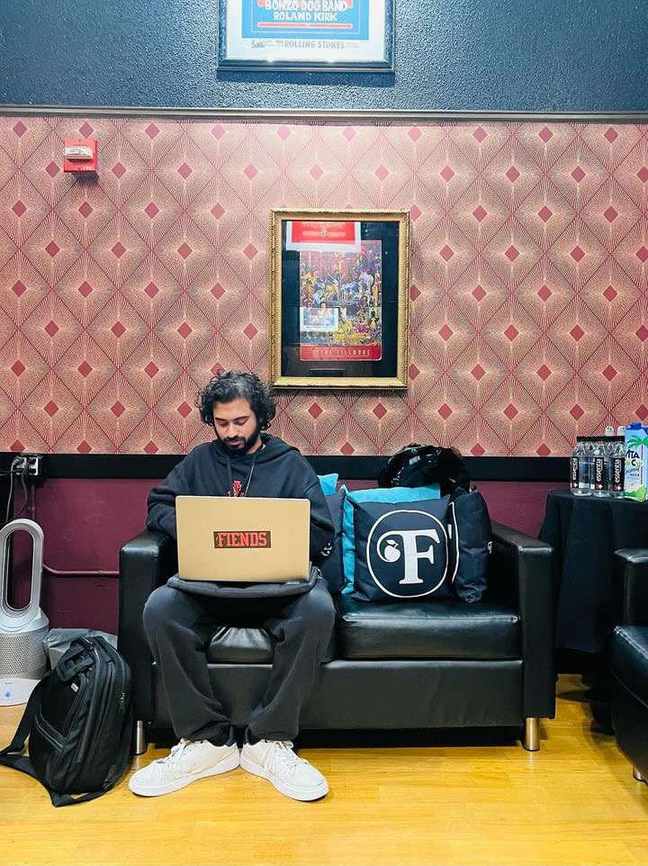 Man with curly hair and beard sitting on a black sofa using a laptop, with bags and water bottles nearby, in a room with patterned wallpaper.