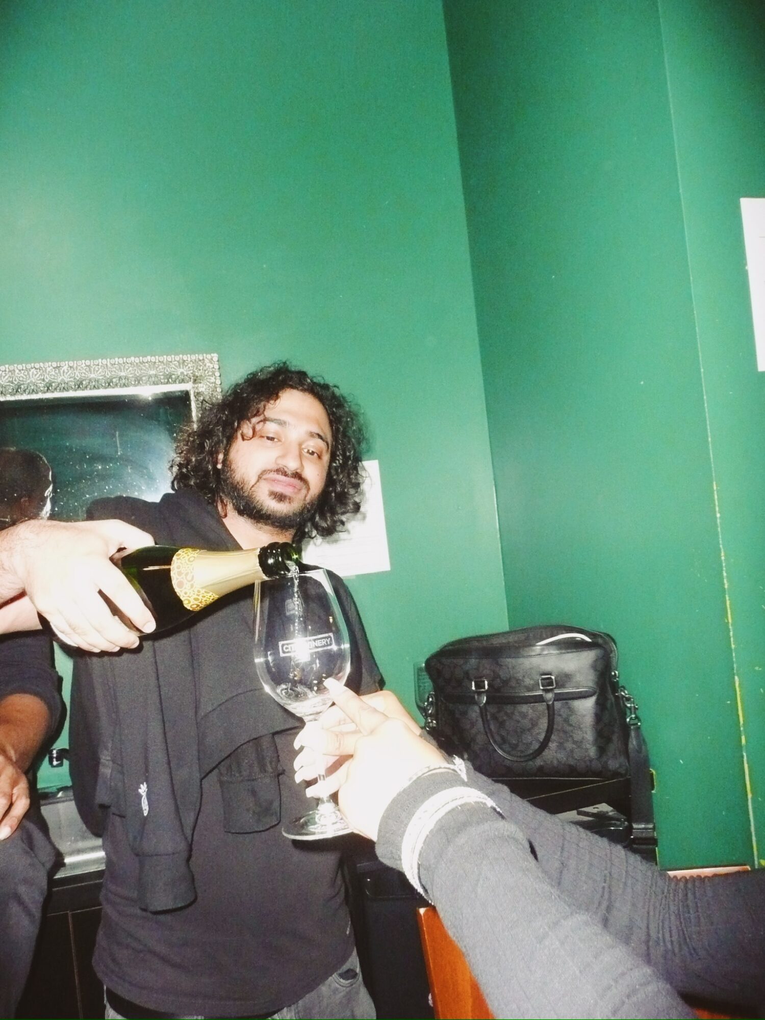 Person with curly hair pours champagne into a glass held by another person, green wall background.