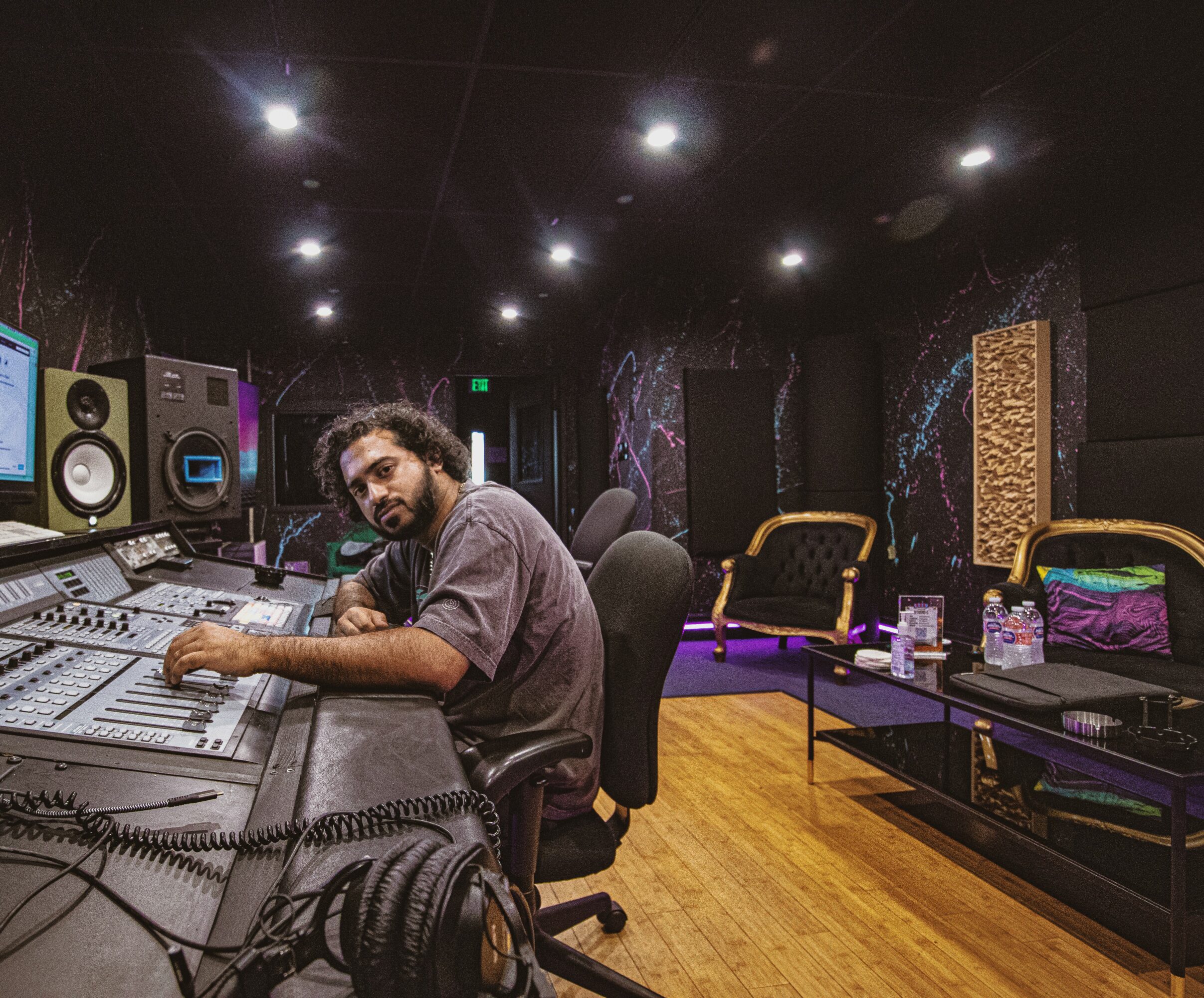 Man with curly hair and beard in a recording studio, sitting at mixing console with equipment and chairs in background.