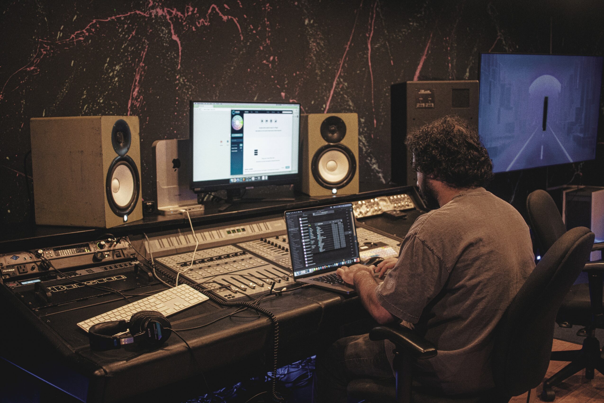 Person working at a music production desk with computer, speakers, and audio equipment in a dark room.