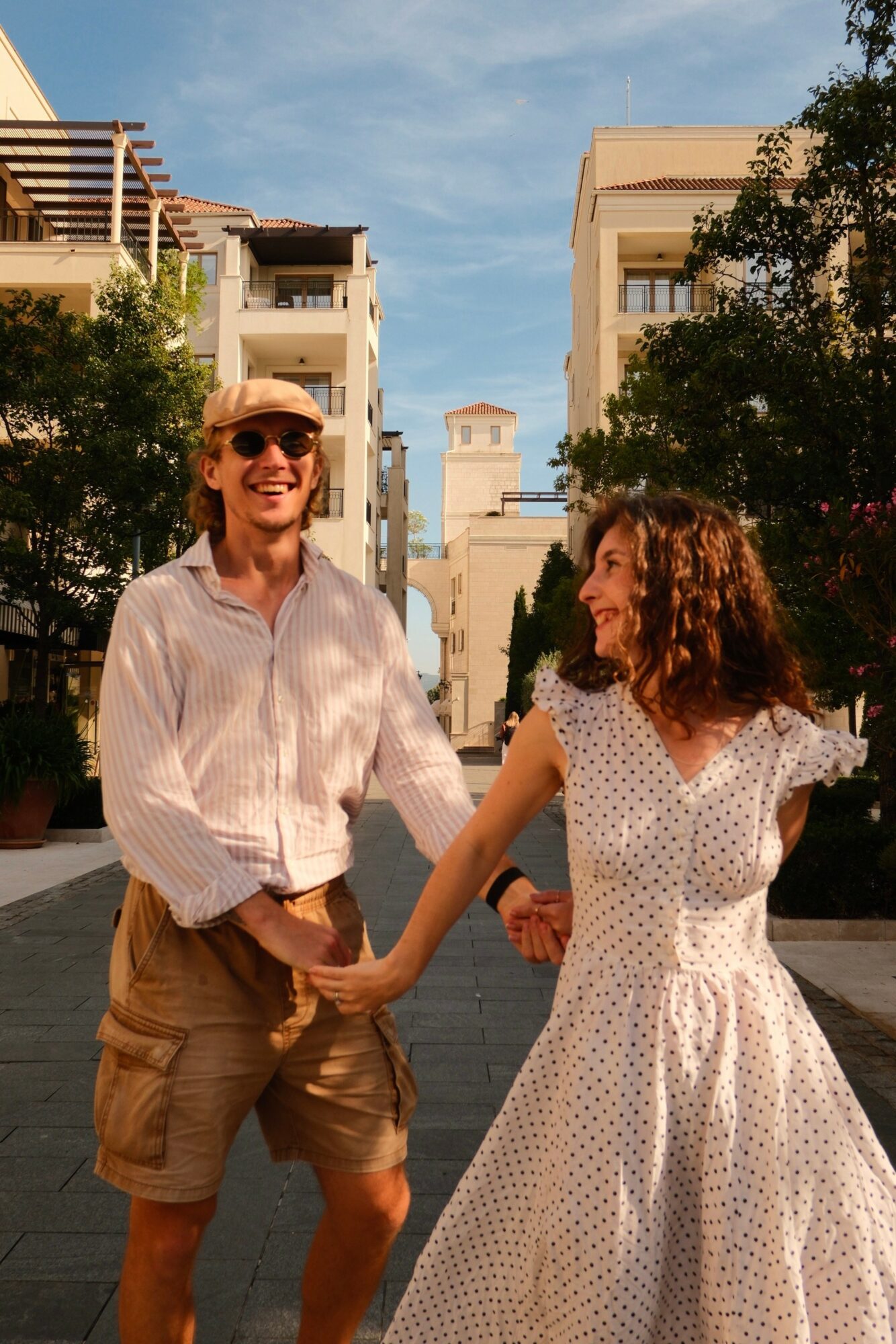 Two people holding hands and smiling outdoors in an urban setting with buildings and trees.
