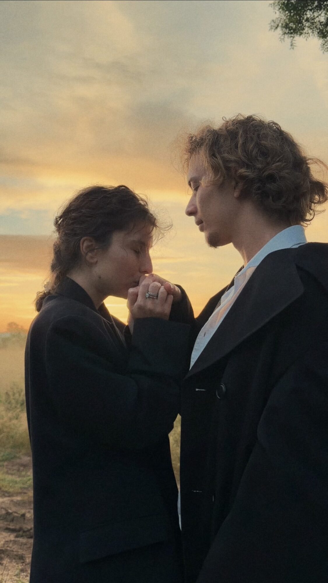 Two people stand close together outdoors during sunset, holding hands and looking at each other.