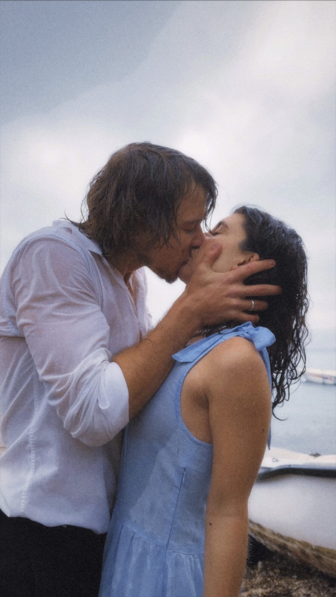 A man and woman kiss outdoors near water, with cloudy sky in background.