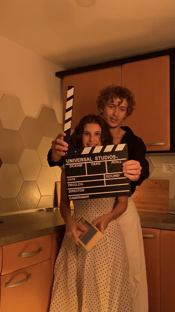 Two people holding a film clapperboard in a kitchen, one with curly hair, the other with straight hair, both smiling.