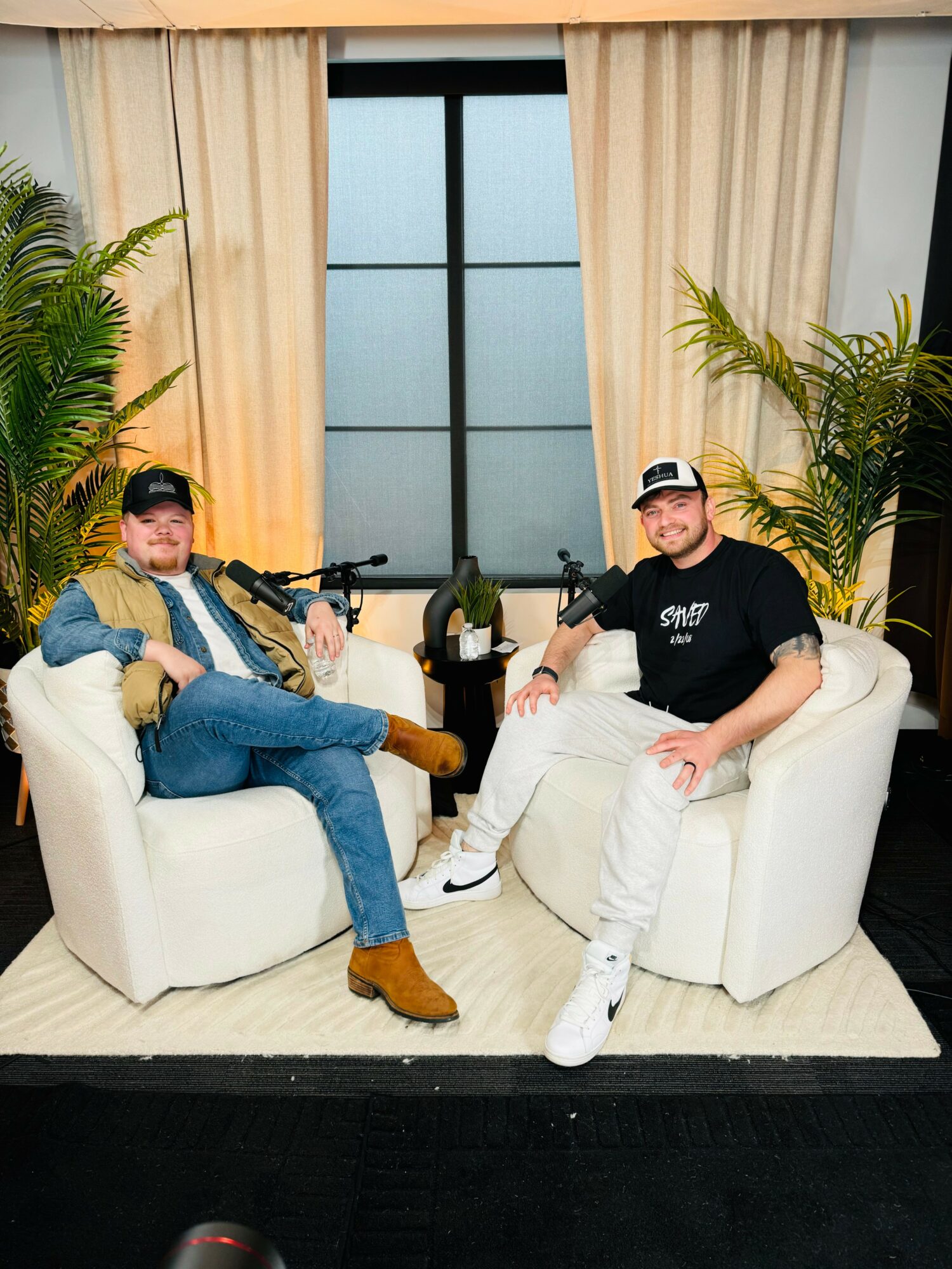 Two men sit on white armchairs in a room with large window, curtains, and potted plants, smiling at the camera.