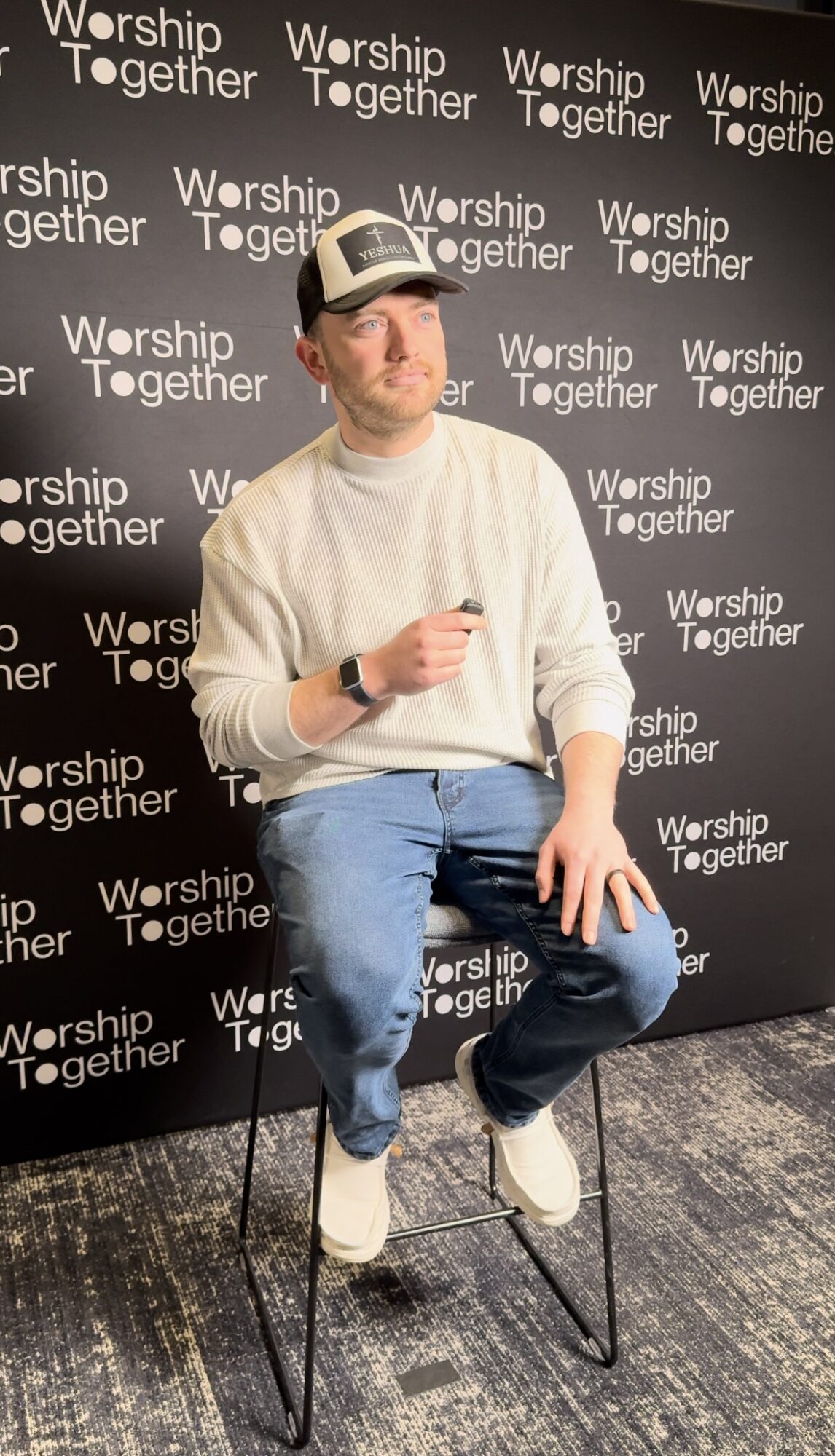 Man sitting on a stool, holding a microphone, in front of a backdrop with 'Worship Together' repeated.