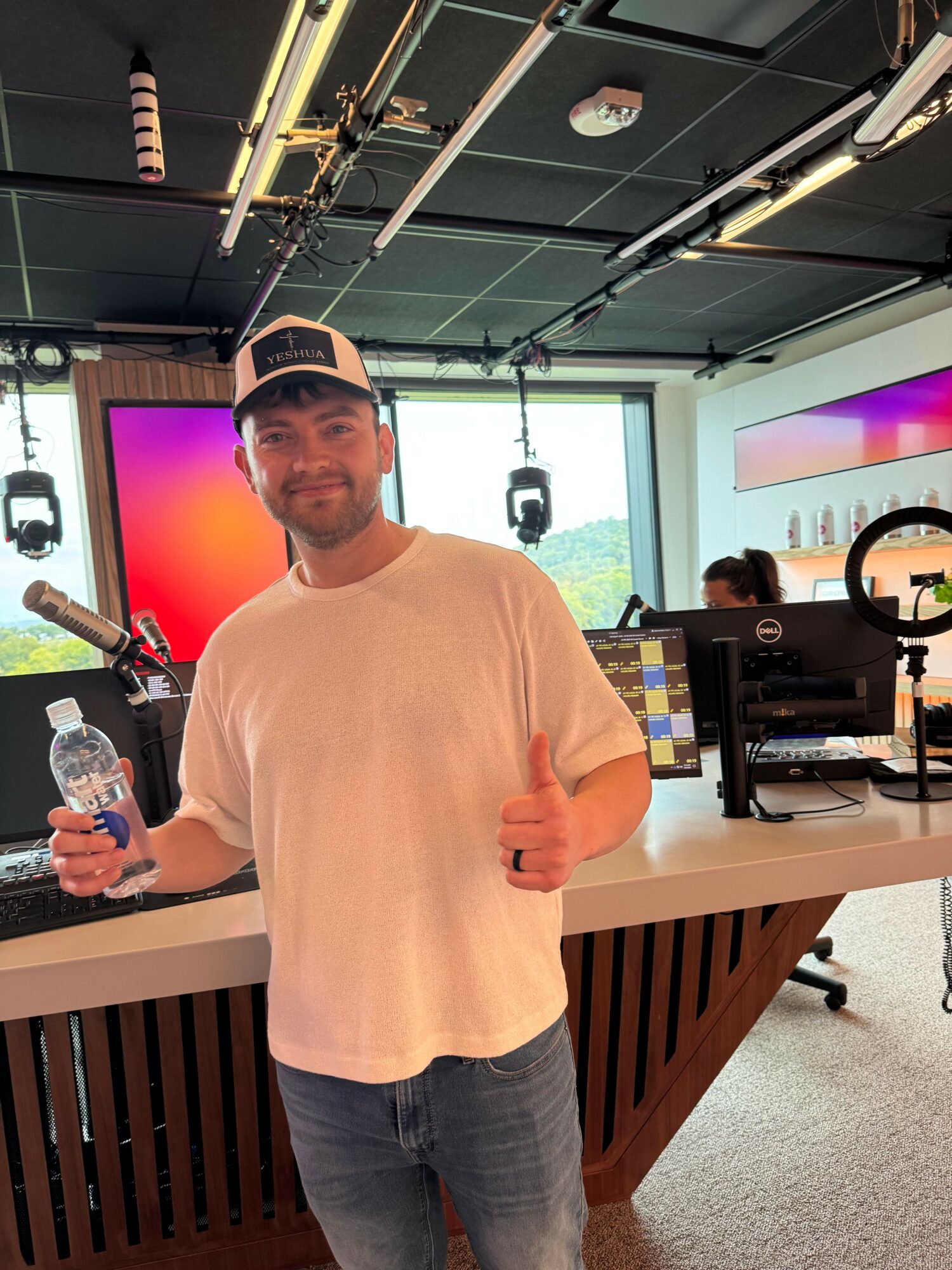 Man in white shirt holding water bottle and giving thumbs up in a modern office with large screen and equipment