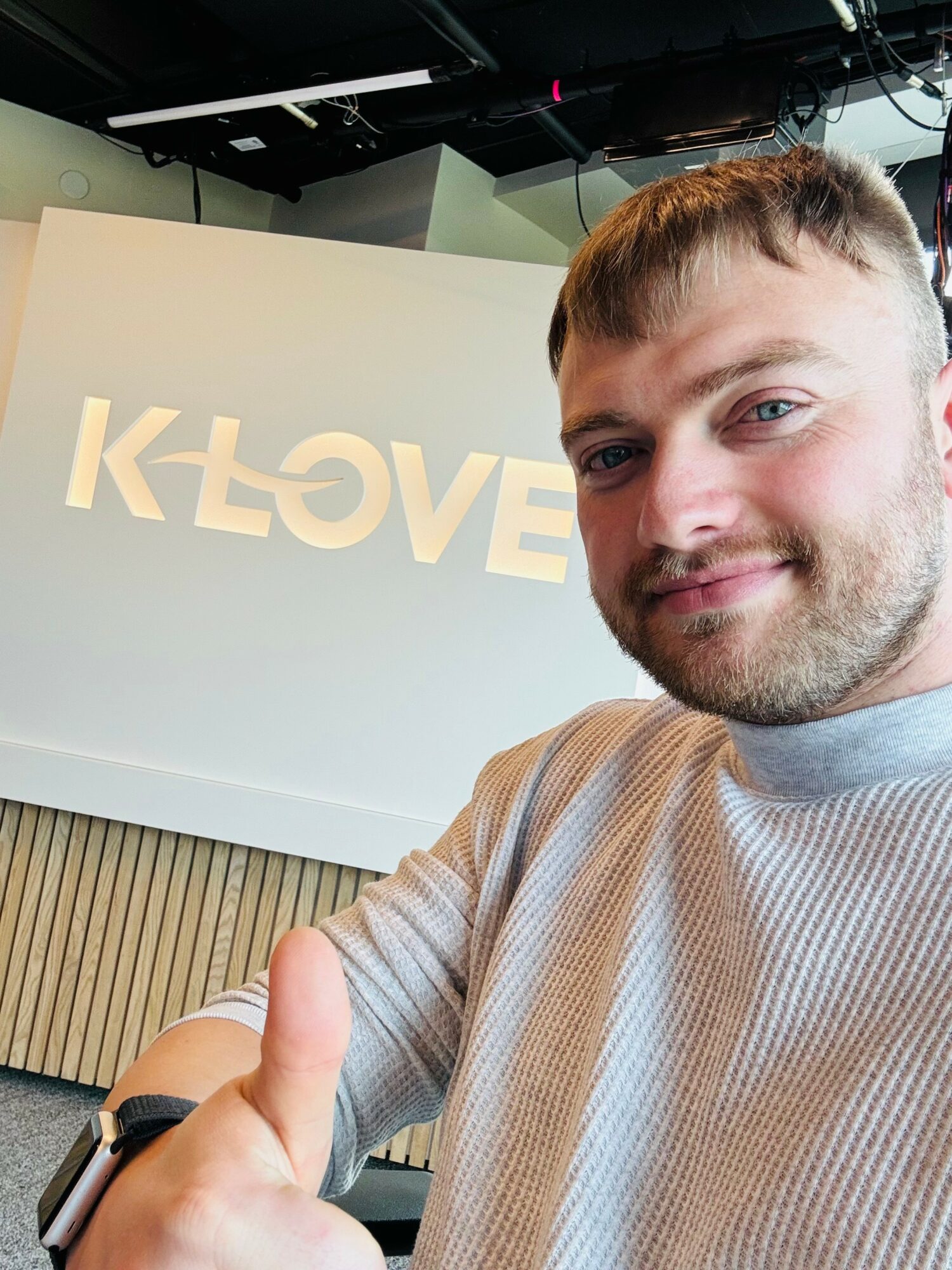 Young man with short hair and beard smiling, giving a thumbs-up, in front of a white wall with a logo.