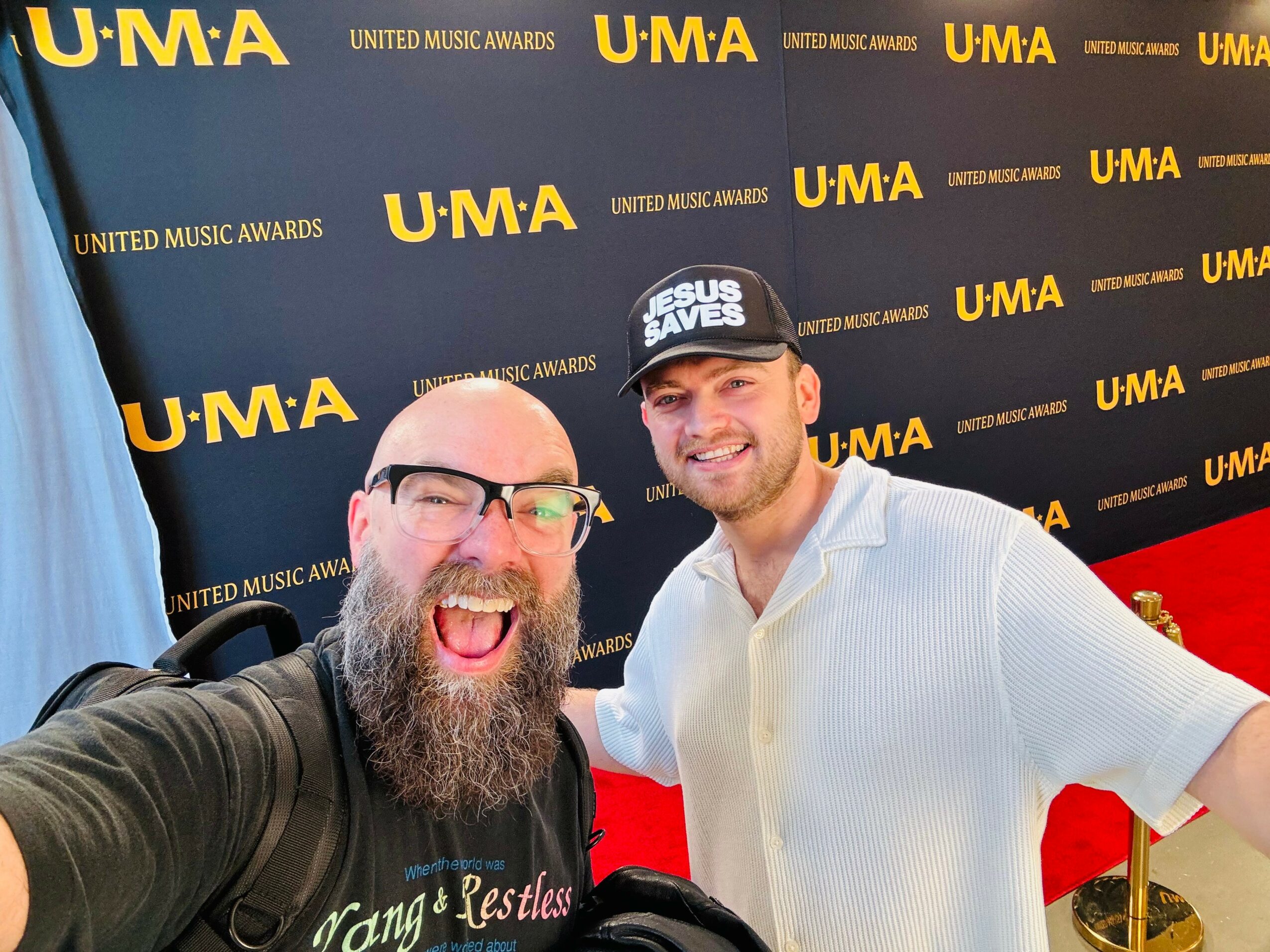 Two men smiling and posing for a selfie at the United Music Awards event, with a black and yellow U.M.A. backdrop.