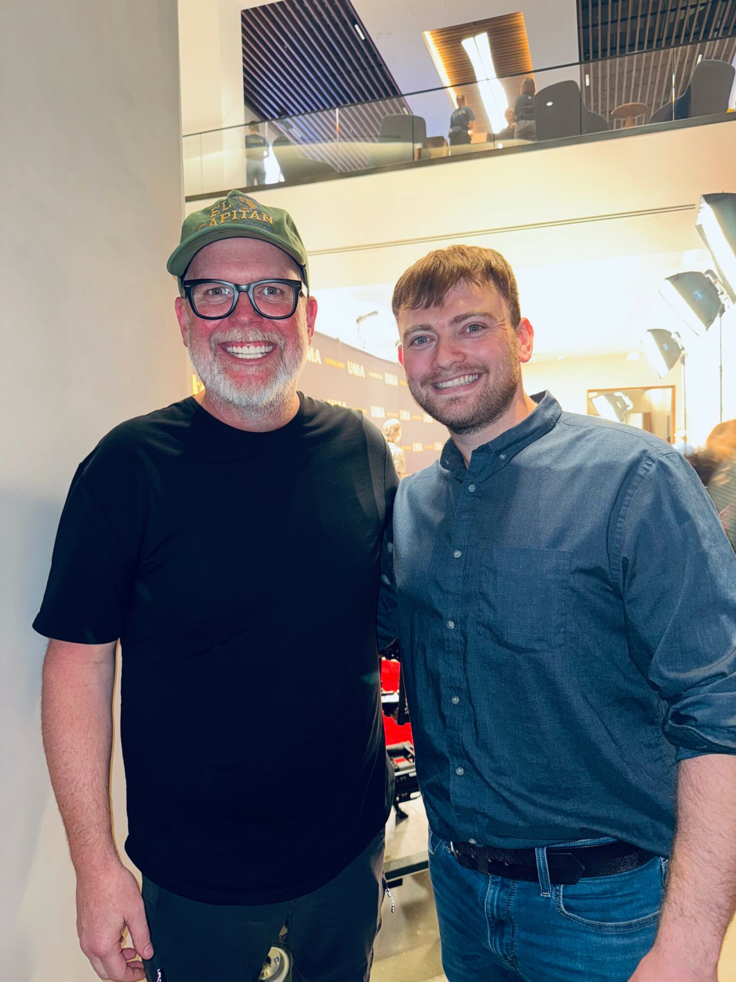 Two men smiling indoors, one wearing glasses and a cap, the other with a beard and a button-up shirt.