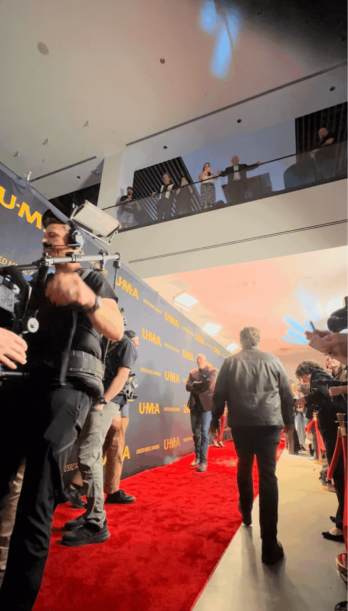People walking on a red carpet at an indoor event with a two-story structure and a blue backdrop.