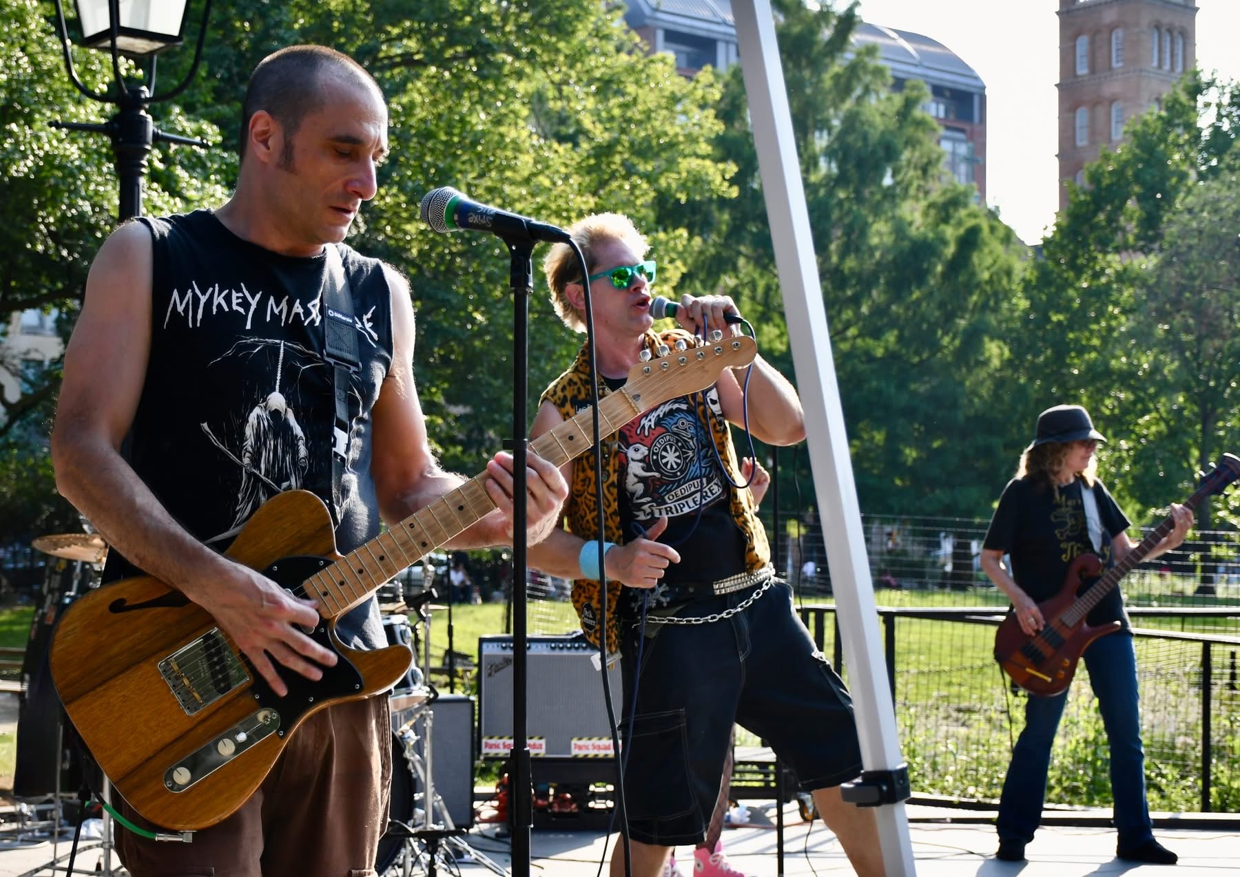Three musicians perform outdoors with trees and buildings in the background, playing guitars and singing into microphones.