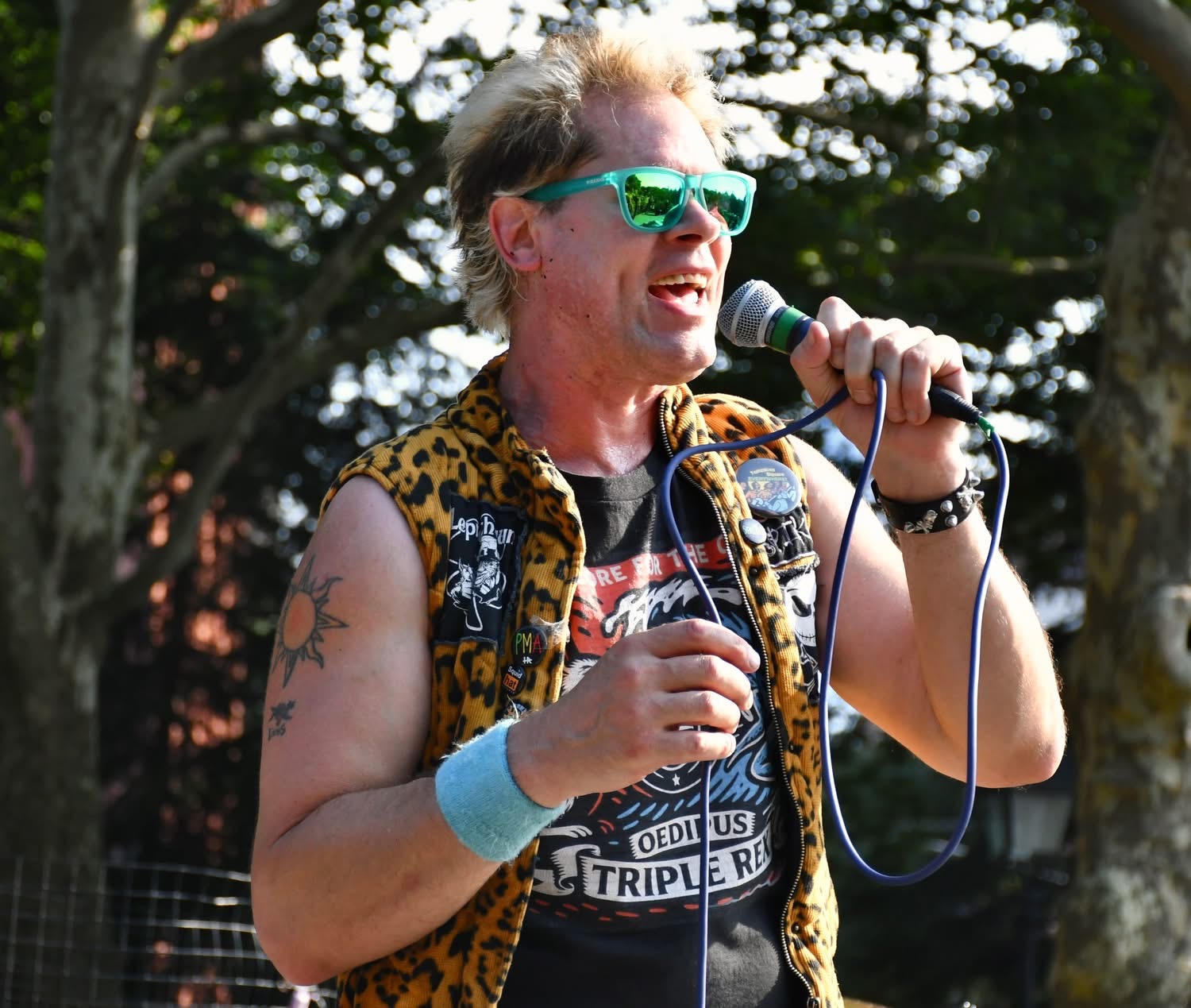 Person with sunglasses speaking into a microphone outdoors, wearing a leopard print vest and a graphic t-shirt.
