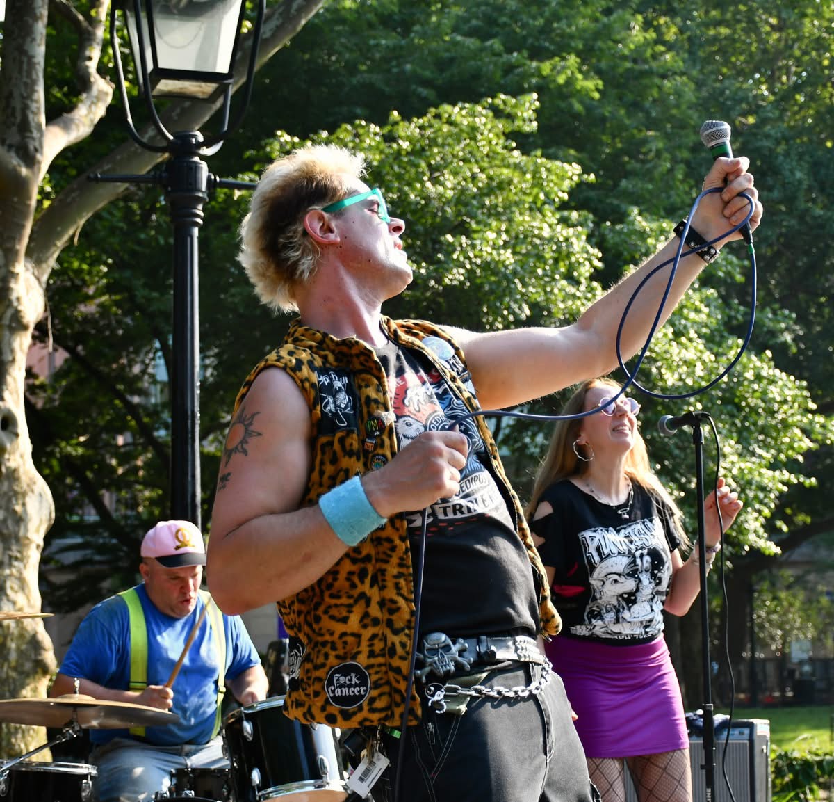 A woman singing into a microphone outdoors with a band, trees, and sunlight in the background.