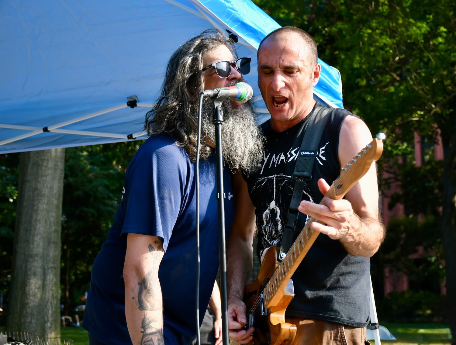 Two men sing and play guitar outdoors under a blue canopy, with trees and sunlight in the background.