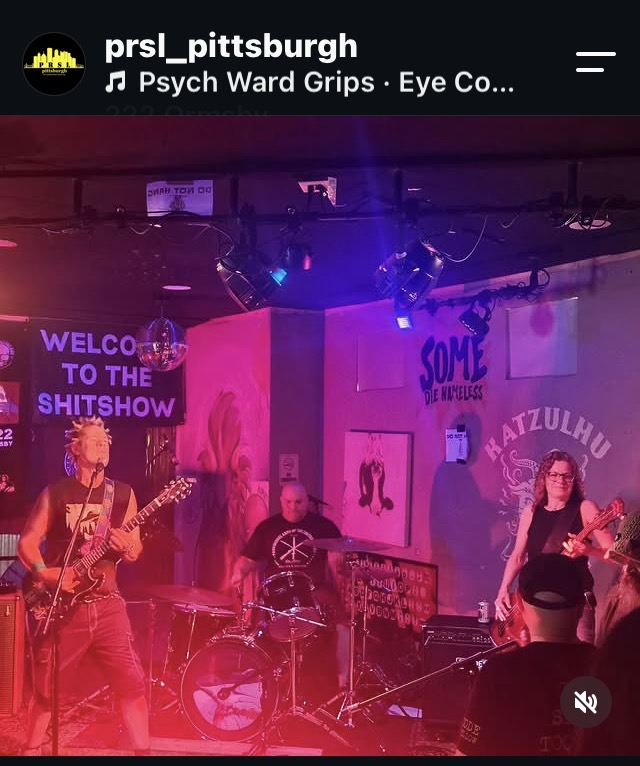 Band performs on stage with pink and purple lighting, musical instruments, and posters in the background.
