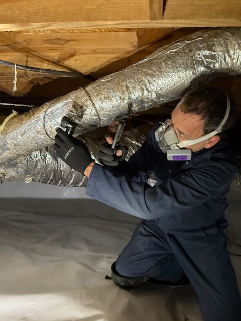Person in protective gear working on a large metallic pipe, lying on the floor, holding a tool or device.