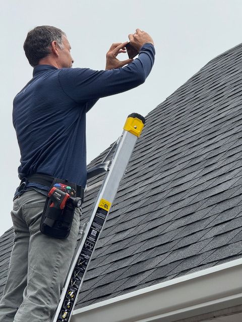 Man standing on a rooftop with a ladder, wearing a blue shirt and gray pants, looking at the sky.
