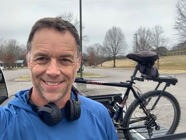 Man smiling outdoors with a bicycle and trees in the background.