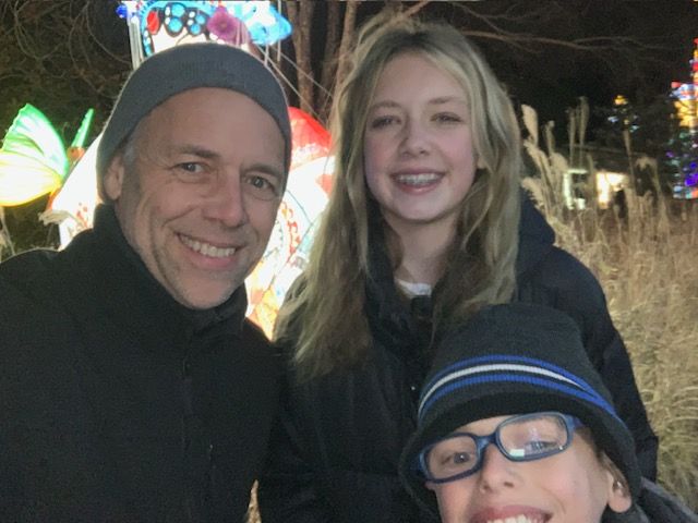 Two people smiling, one wearing glasses and a cap, the other with long hair, outdoors at night with lights in background.