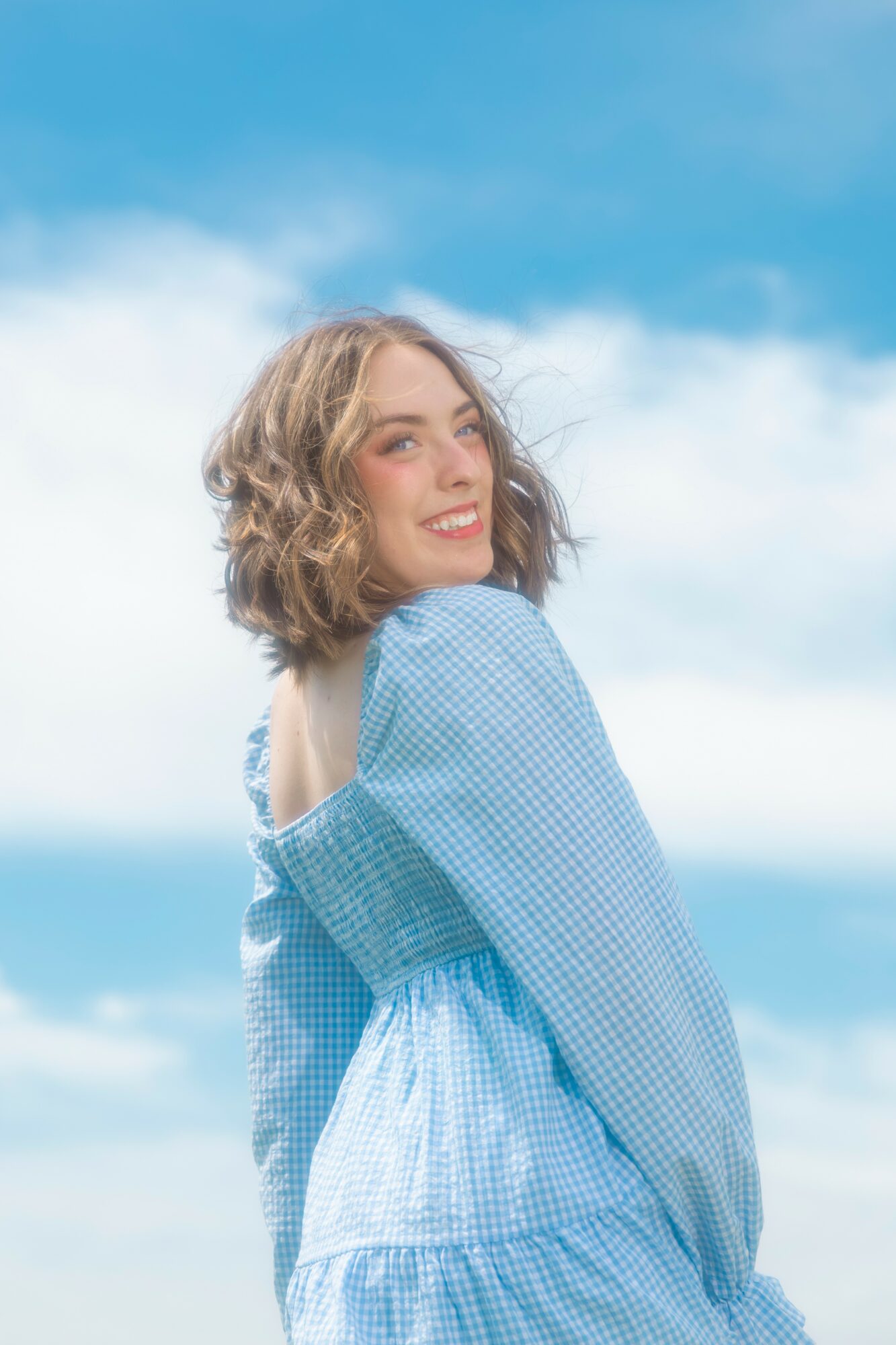 Young woman with curly hair smiling outdoors against a blue sky with clouds, wearing a blue dress.