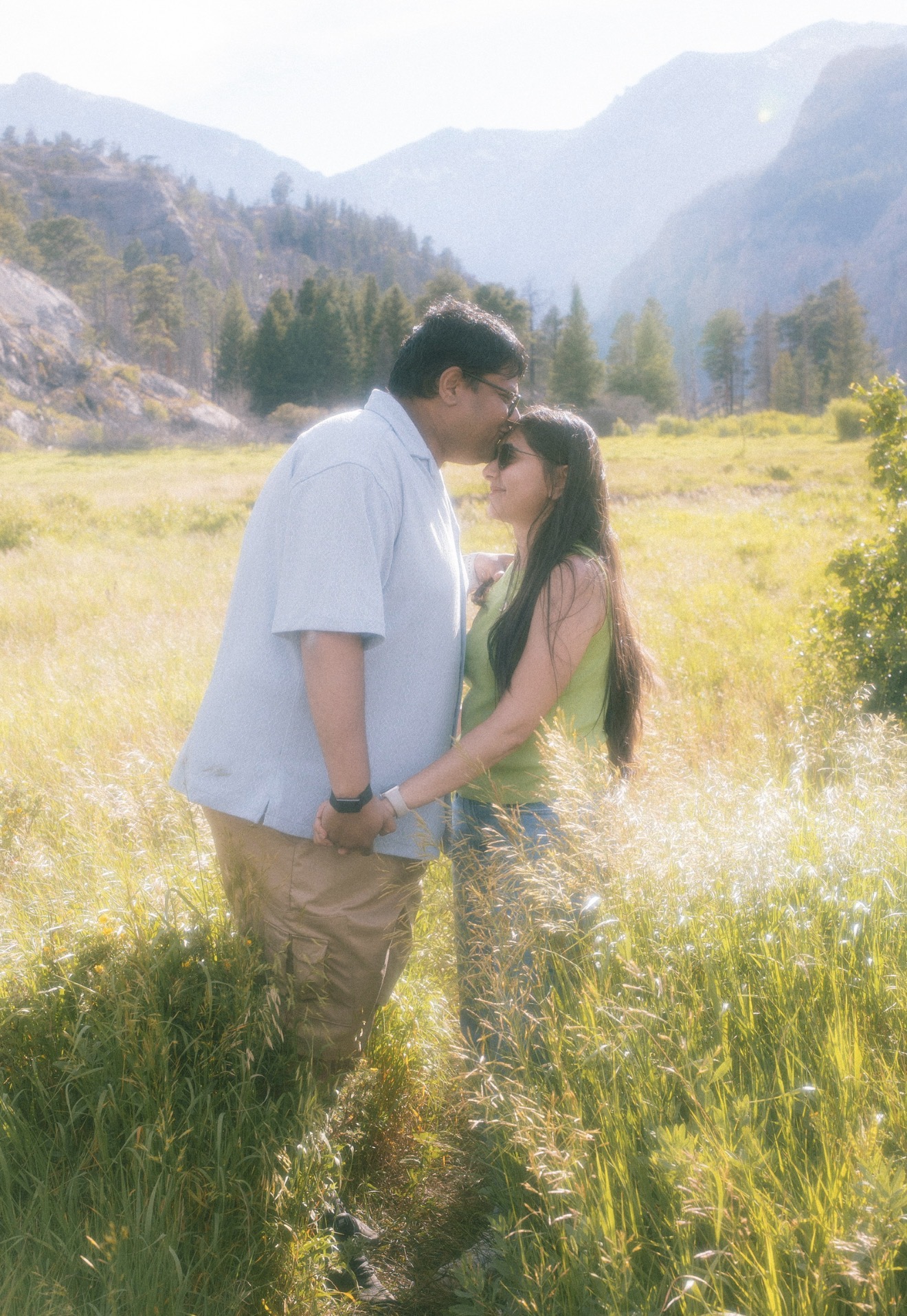 A couple stands close in a grassy field with mountains and trees in the background, holding hands and looking at each other.