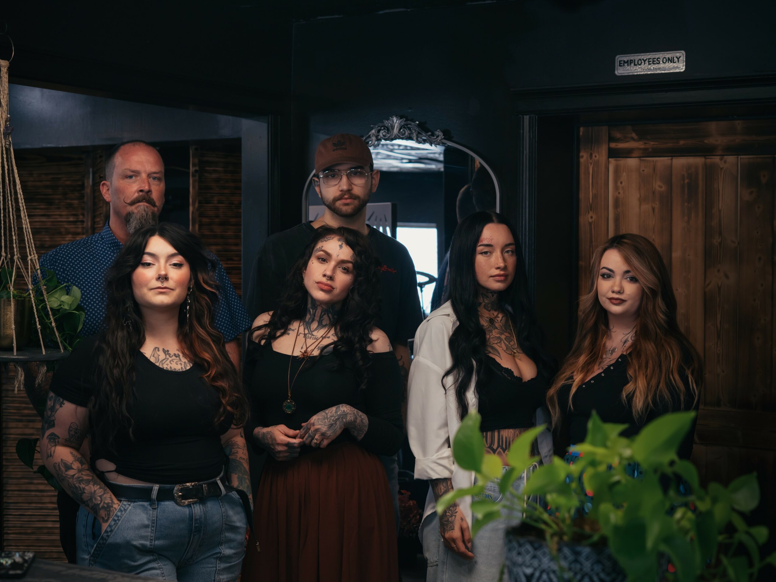 Group of six people standing indoors, some with tattoos, in a dimly lit room with wooden and brick decor.