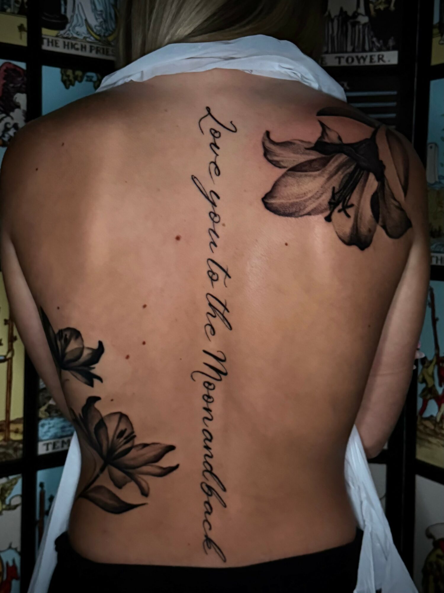 Back of a person with a tattoo of flowers and a quote, with a tattooed shoulder and a white top hanging off one shoulder.