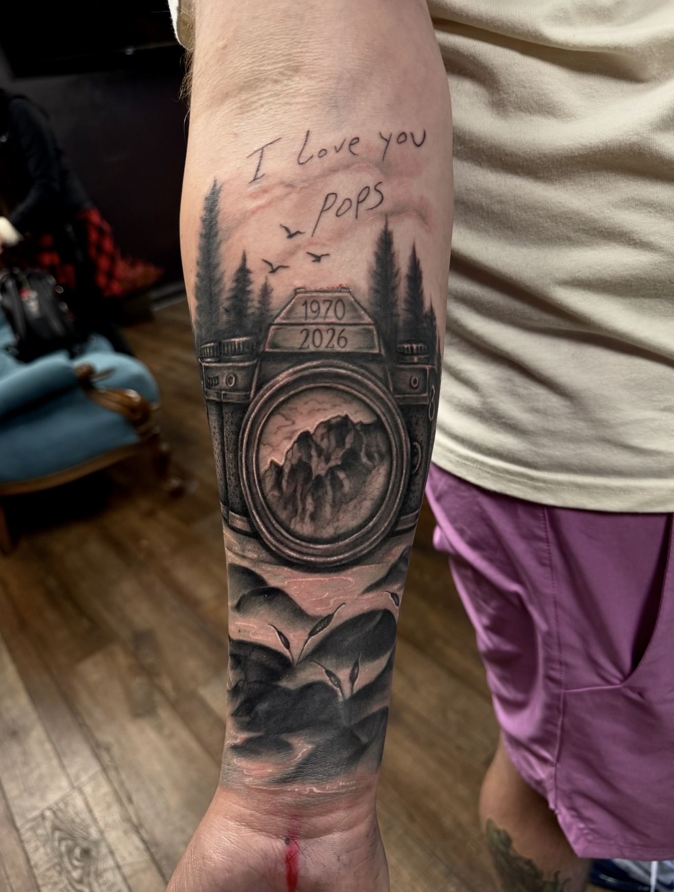 Tattoo of a camera with mountains inside, trees, birds, and the words 'I love you Pops' and years 1970-2026, on a person's arm.