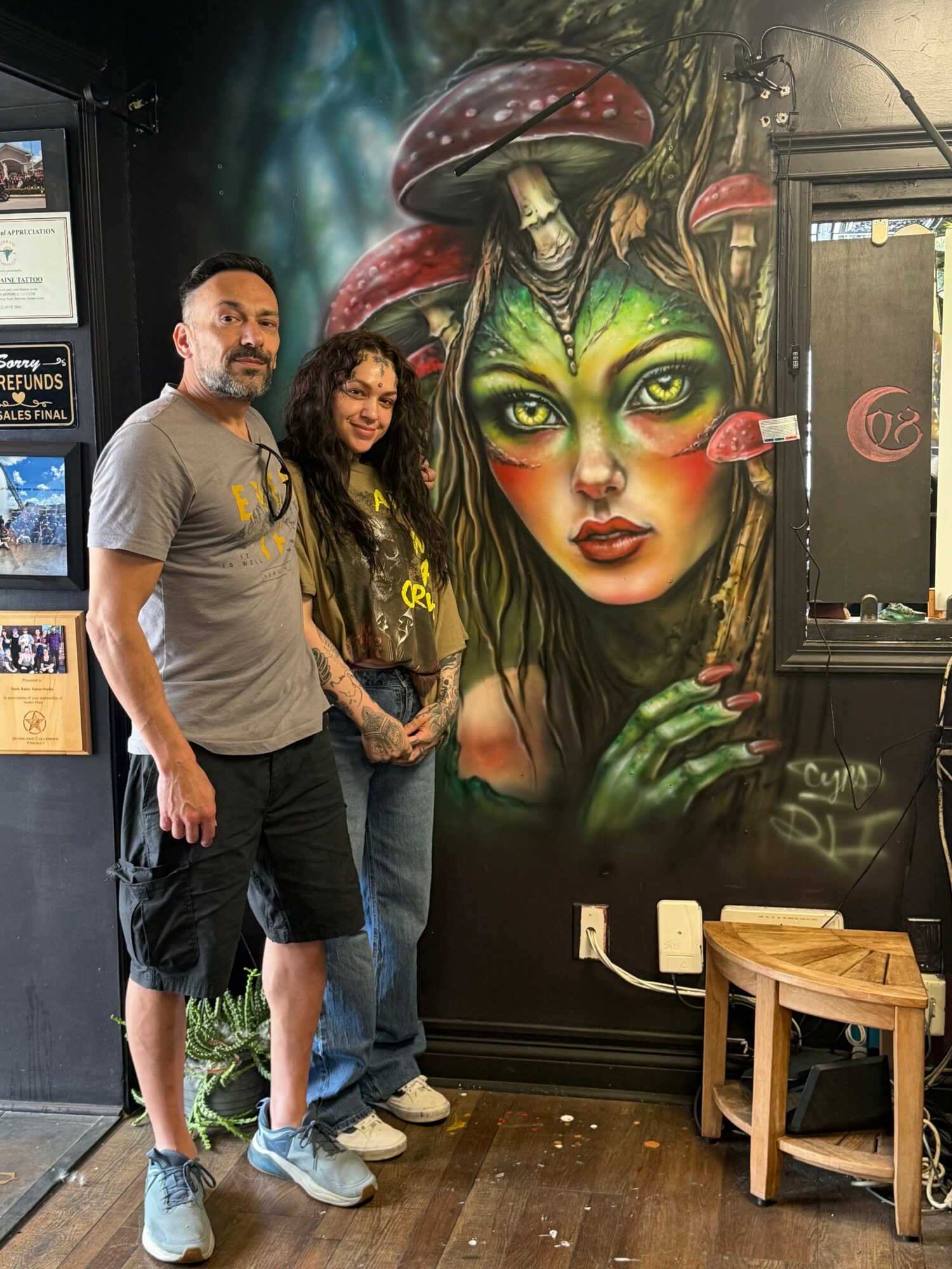 Two people stand in front of a colorful mural of a woman with green skin, mushrooms, and plants, inside a cafe or shop.