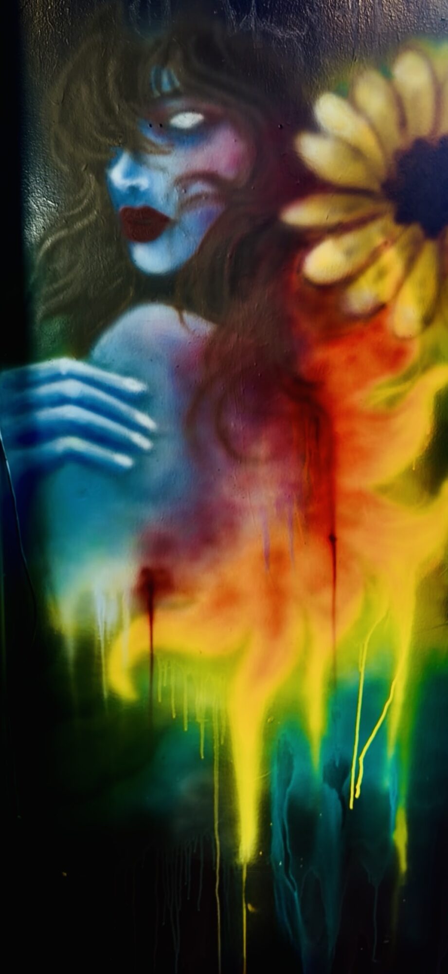 A painted portrait of a woman with blue skin, dark hair, and red lips, surrounded by a sunflower and colorful abstract elements.