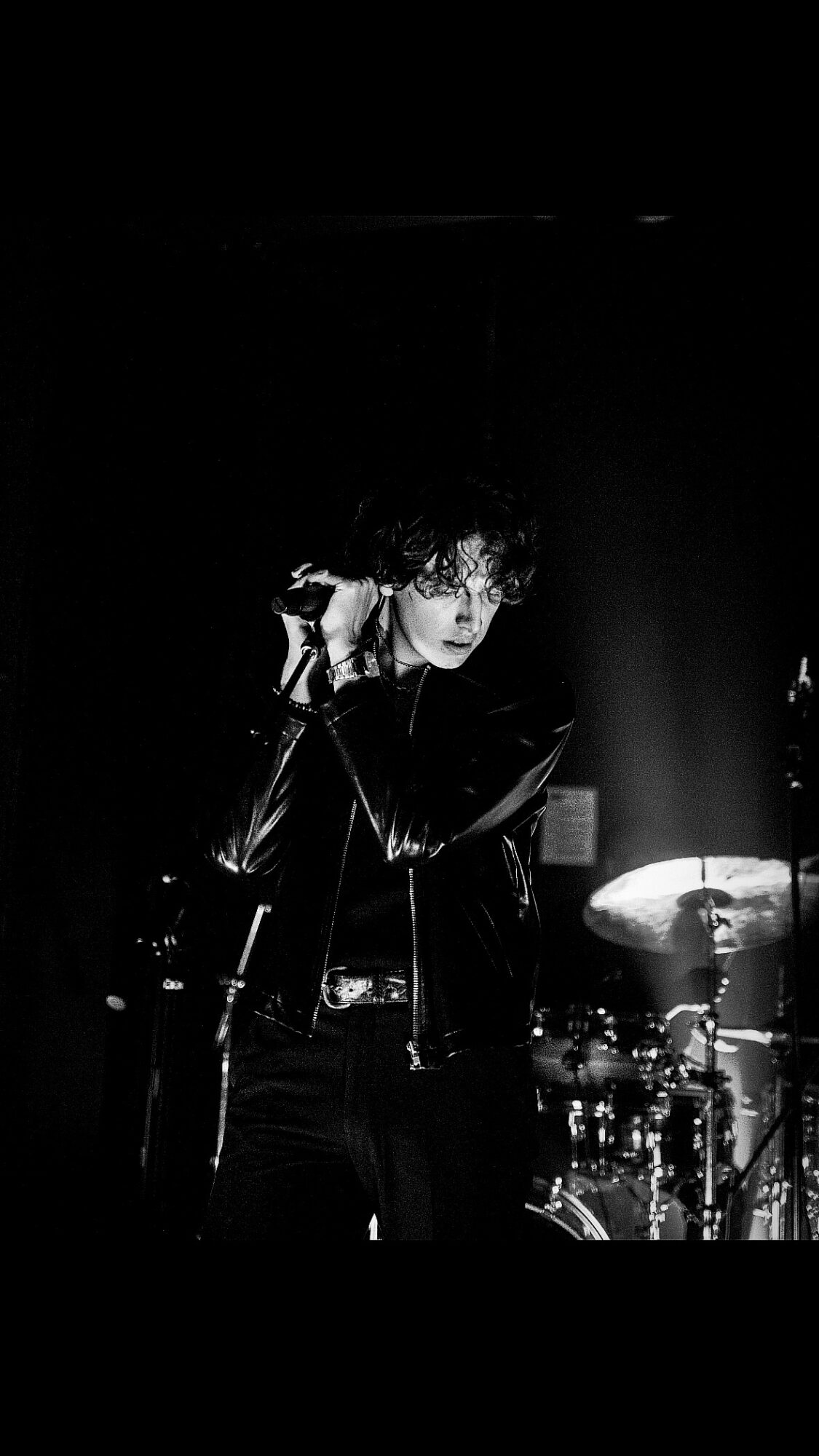 Person with curly hair singing into a microphone on stage, with drums in the background, in black and white.