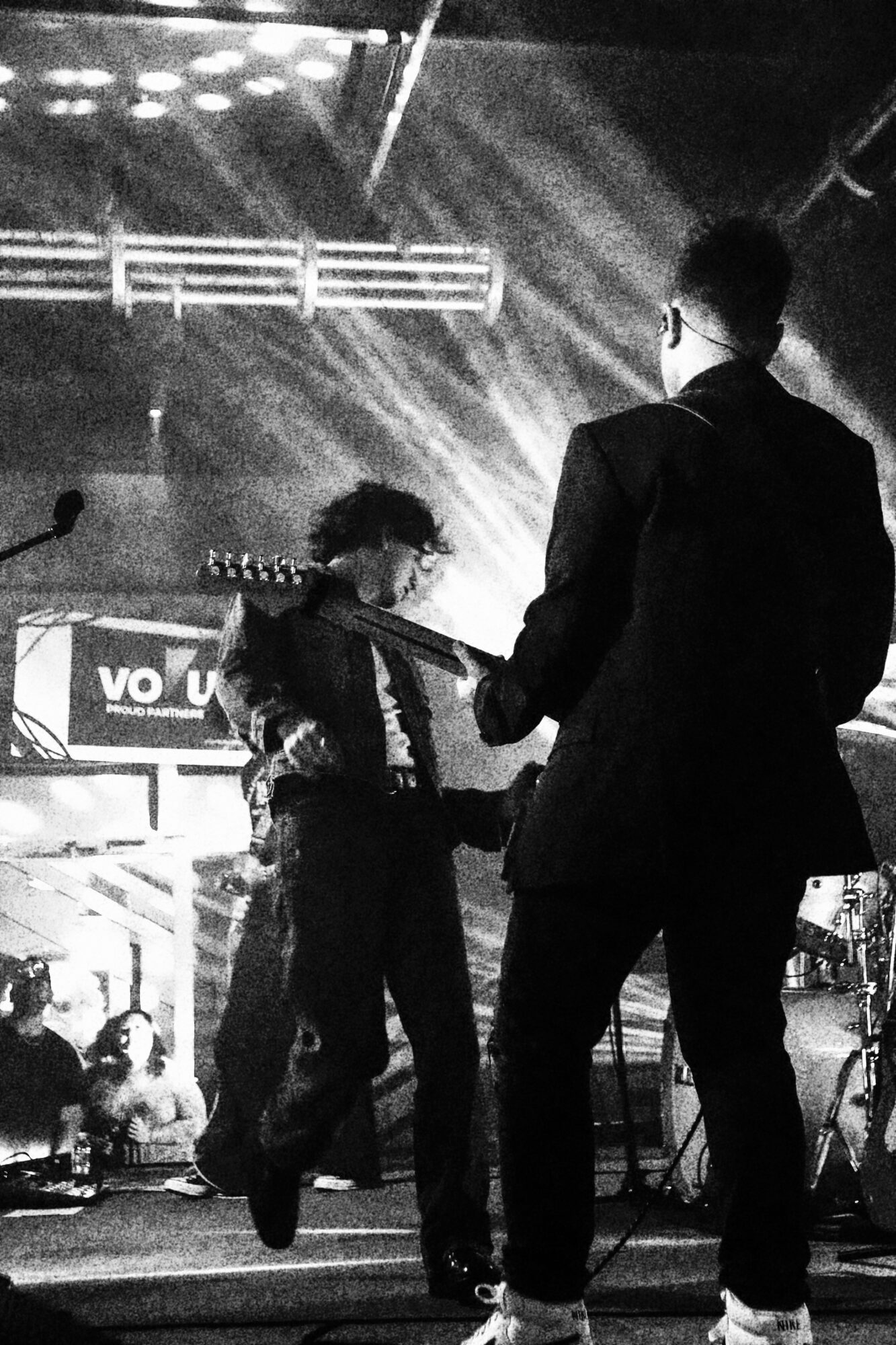 Two musicians perform on stage with bright lights and a crowd in the background.