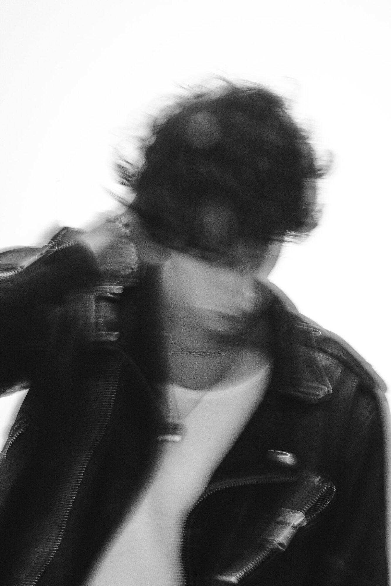 Person with curly hair wearing a leather jacket and white shirt, head tilted downward, blurred face, in black and white.