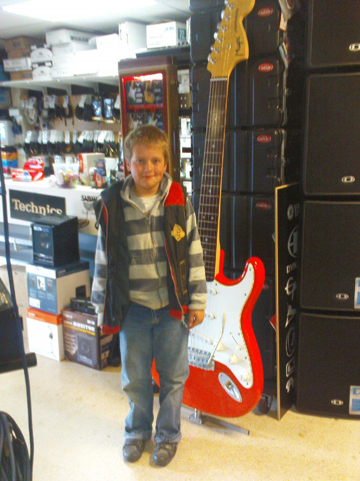 Child standing in a store aisle with musical instrument displays, smiling, wearing a striped shirt and jacket.