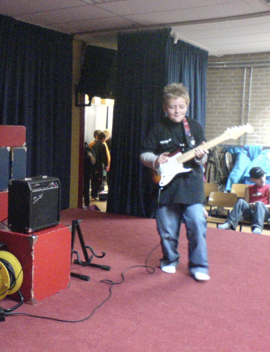 Person playing electric guitar on stage with amplifier, red carpet, and audience in background.