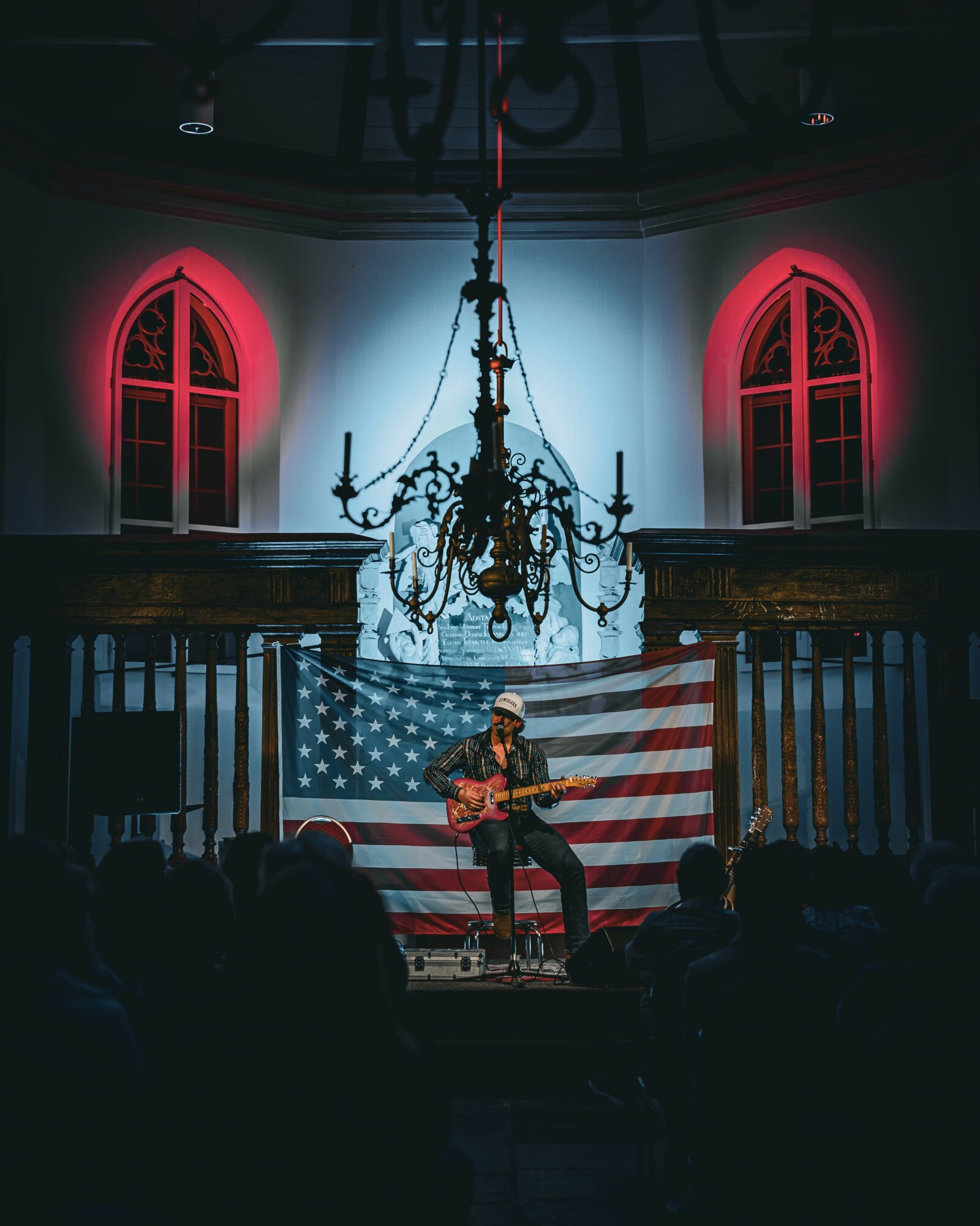 Person playing guitar on stage with American flag backdrop inside a church, audience in foreground, chandelier overhead, red lighting on windows.