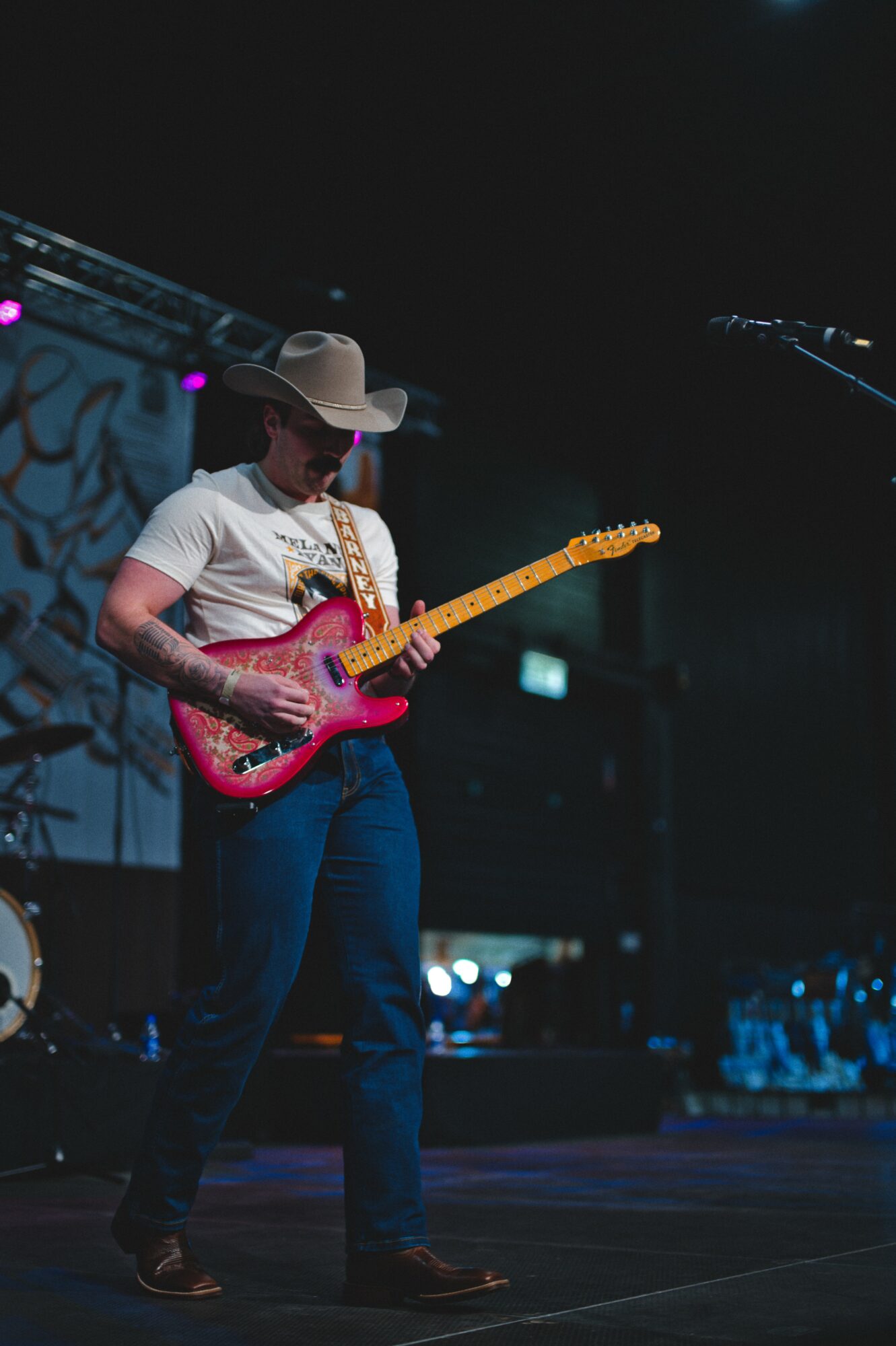 Person playing an electric guitar on stage, wearing a hat and casual clothing, with stage lights in the background.