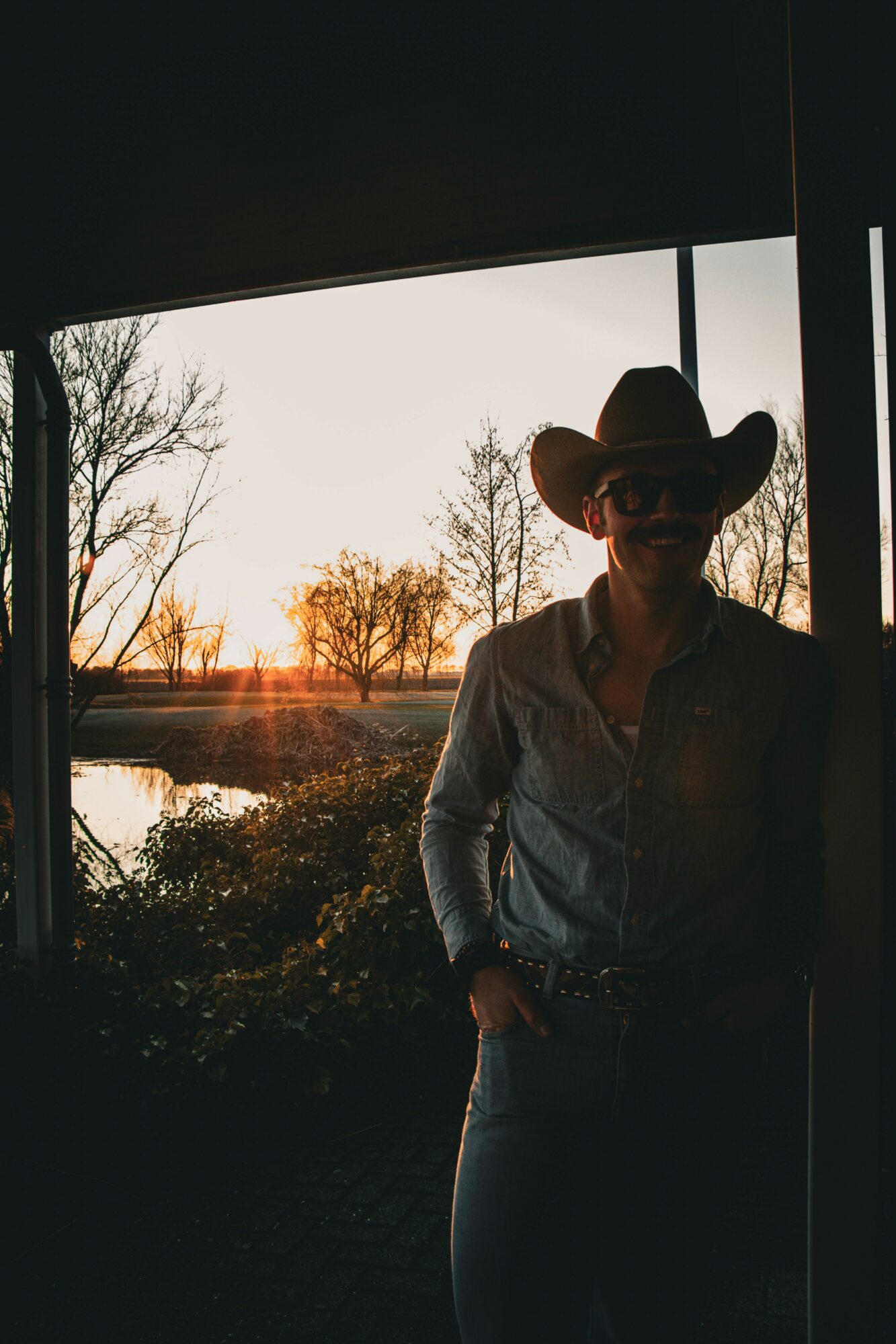 Person wearing a cowboy hat and sunglasses standing outdoors during sunset, trees and a river in background.